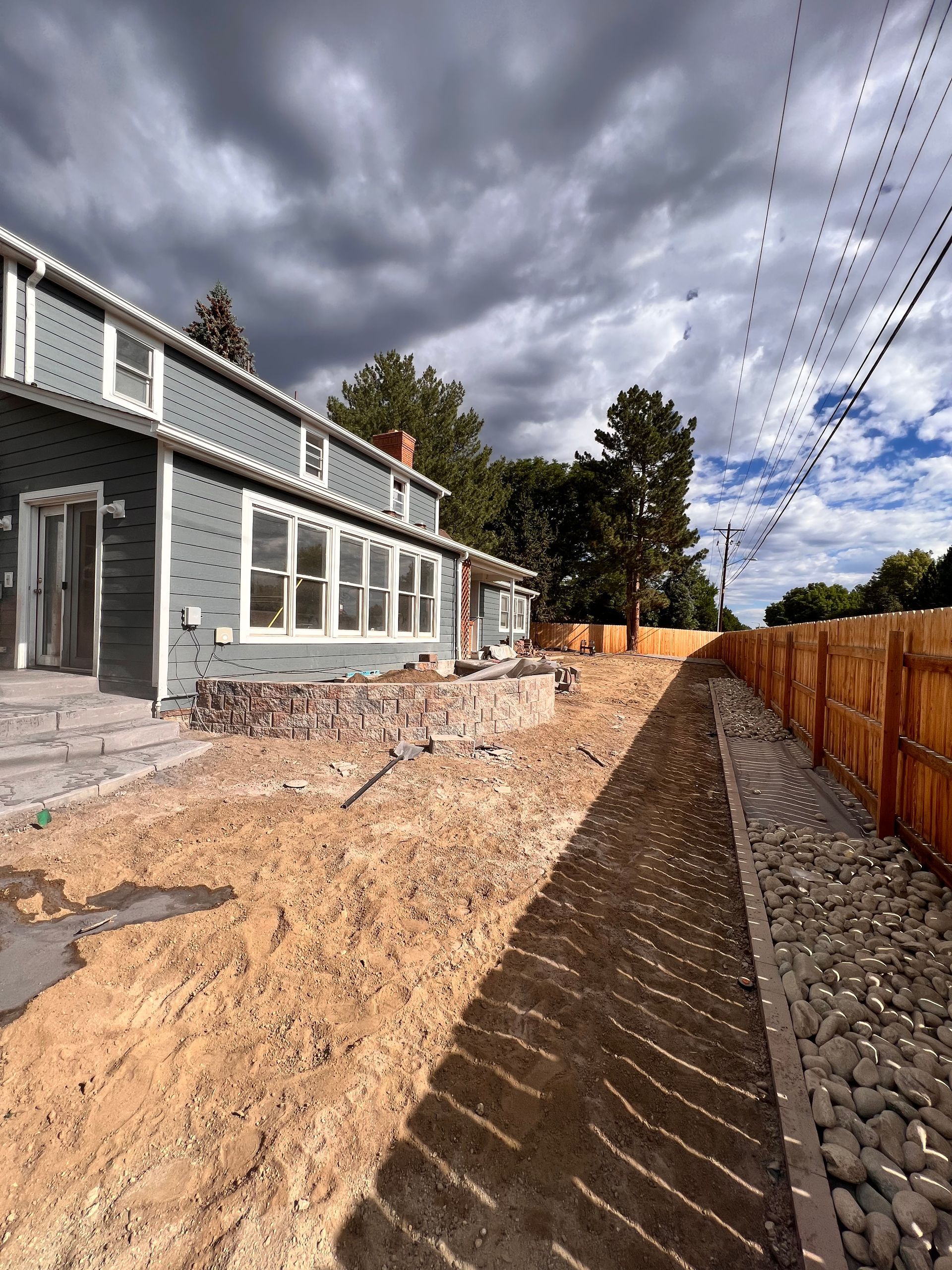 Two-story gray house with unfinished backyard. Brown dirt, wooden fence, cloudy sky.