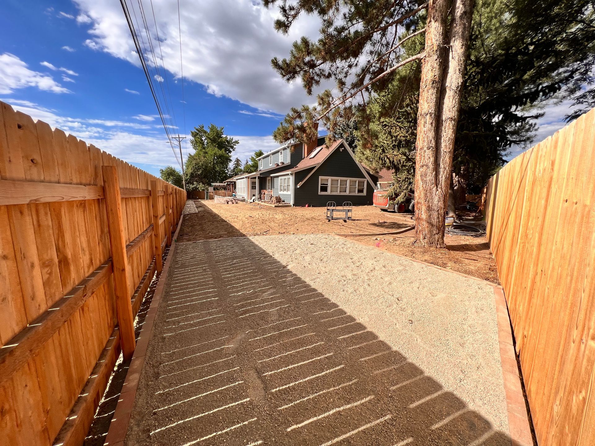 Narrow gravel driveway between wooden fences, leading to a house with trees and blue sky.