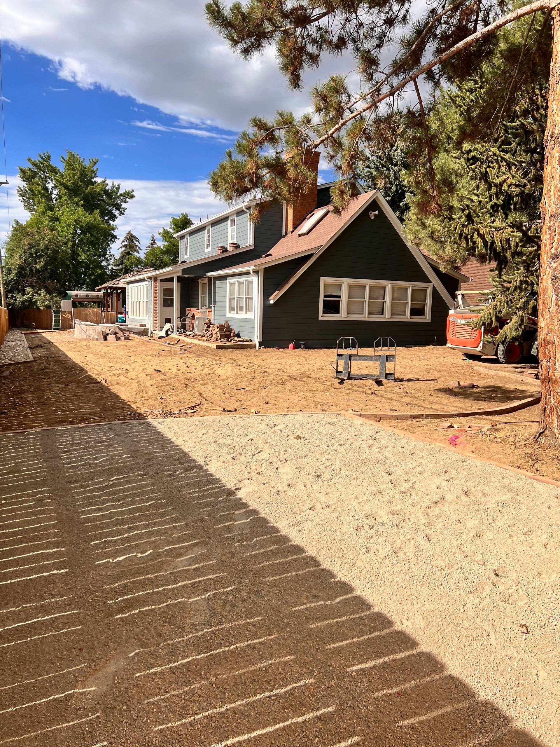 Gravel driveway leads to a two-story house with green siding and a cleared yard under a cloudy sky.