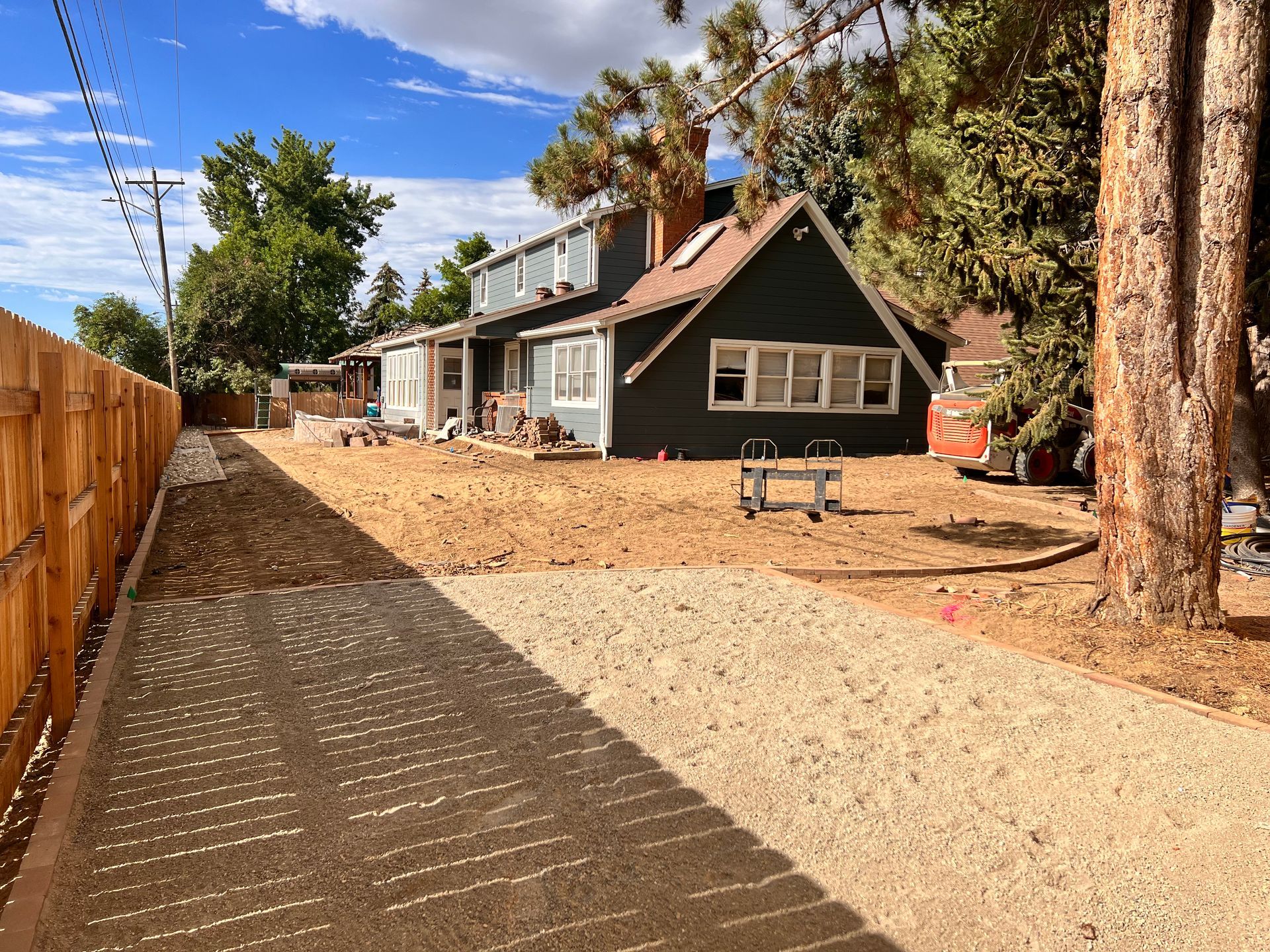 Gravel driveway leads to a dark green house with a wooden fence on the left and trees on the right.