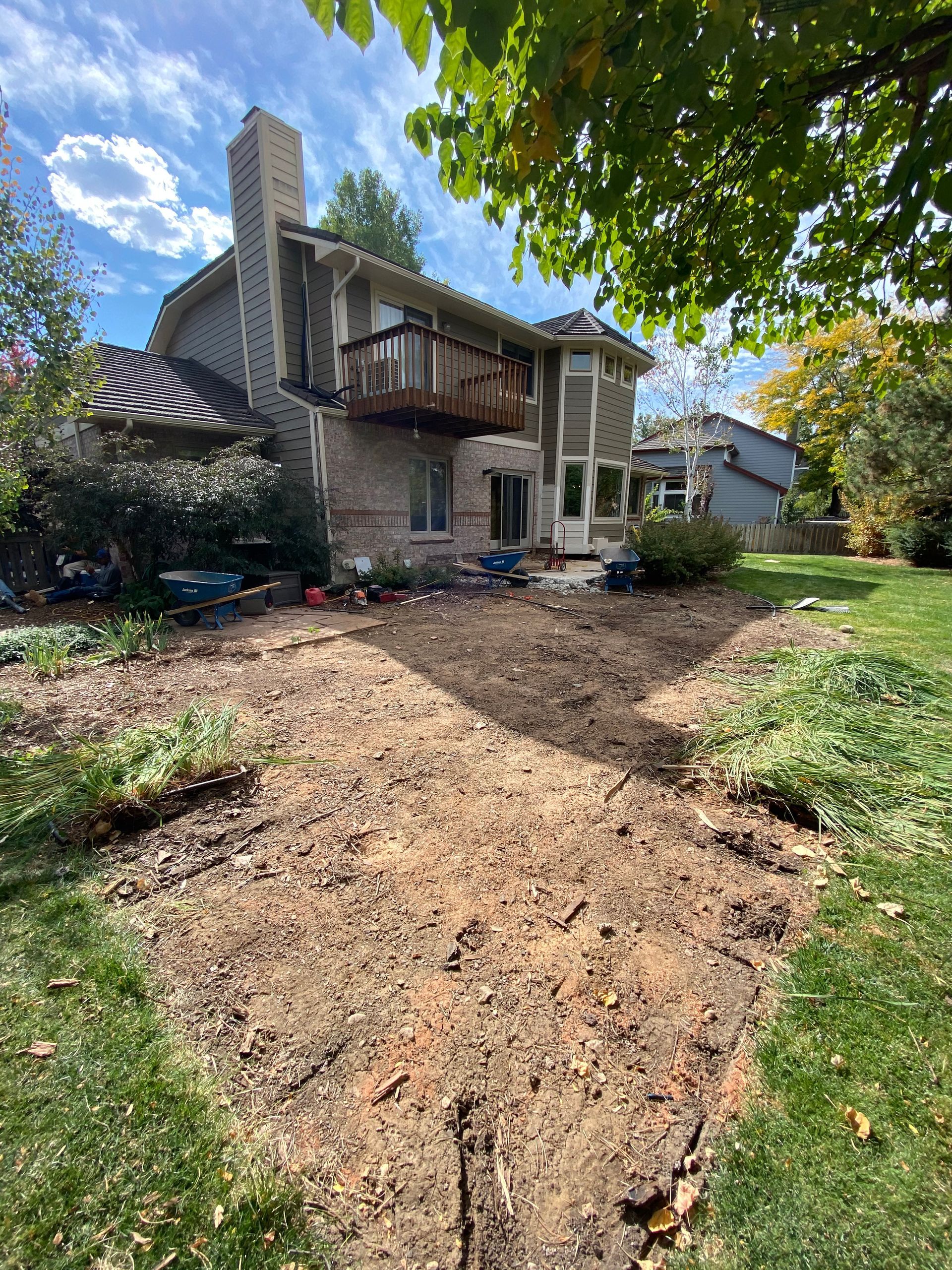 A backyard excavation site with a house, deck, and chimney in the background under a blue sky.