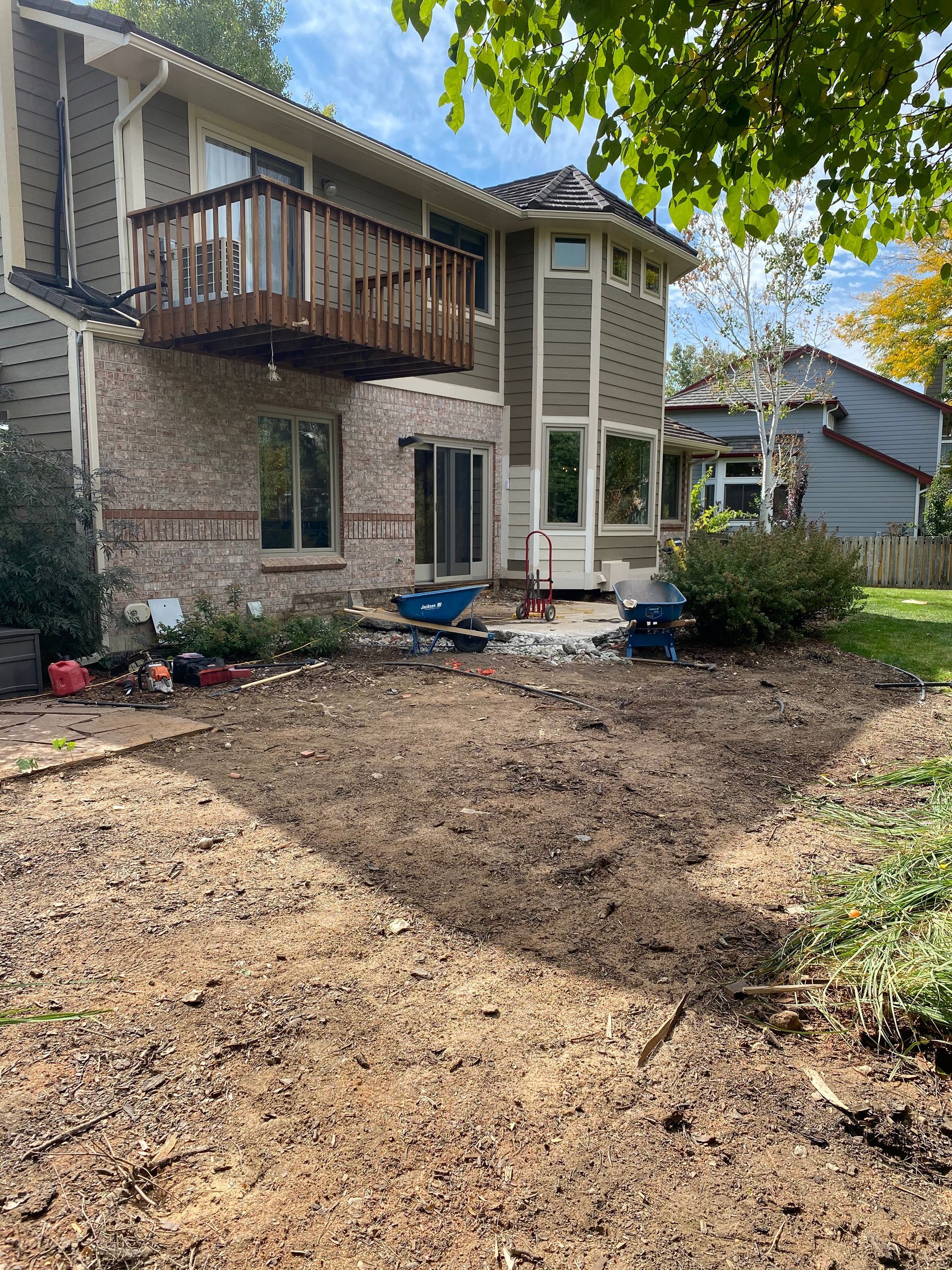 Backyard of a house with a deck. Dirt patch in the foreground, bushes and tools near the house. Sunny day.