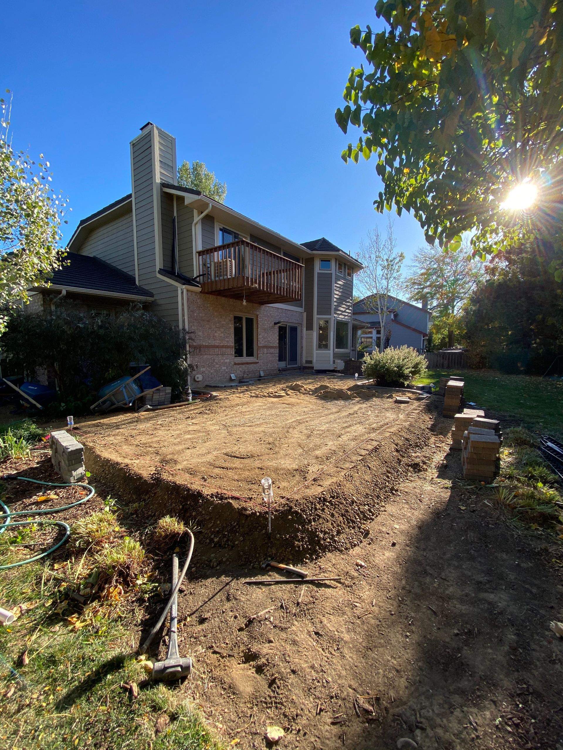 Backyard with excavated ground, awaiting a patio, in front of a two-story brick house with a balcony.