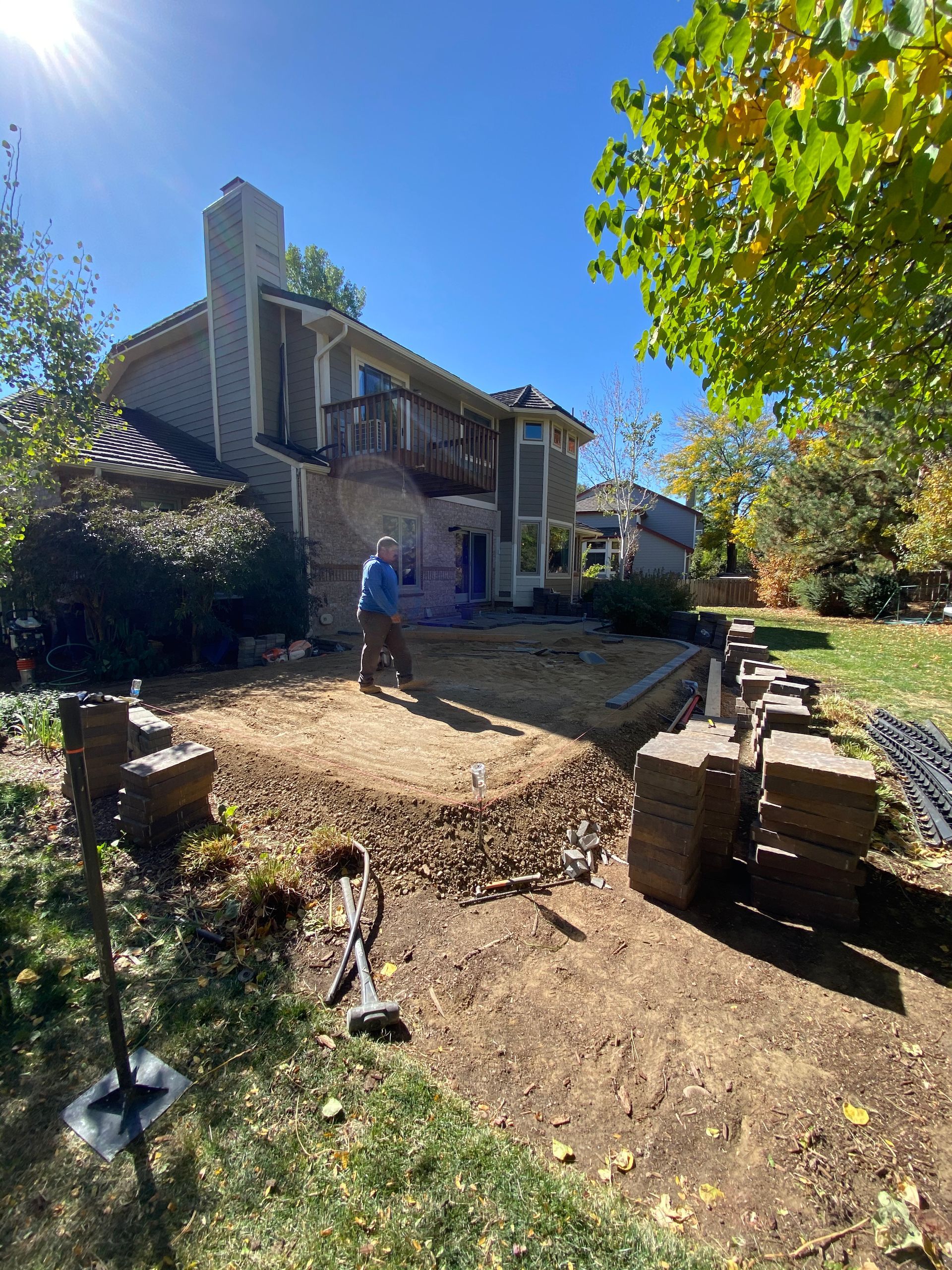 Man working on a backyard patio, brown bricks, house, blue sky, fall foliage.