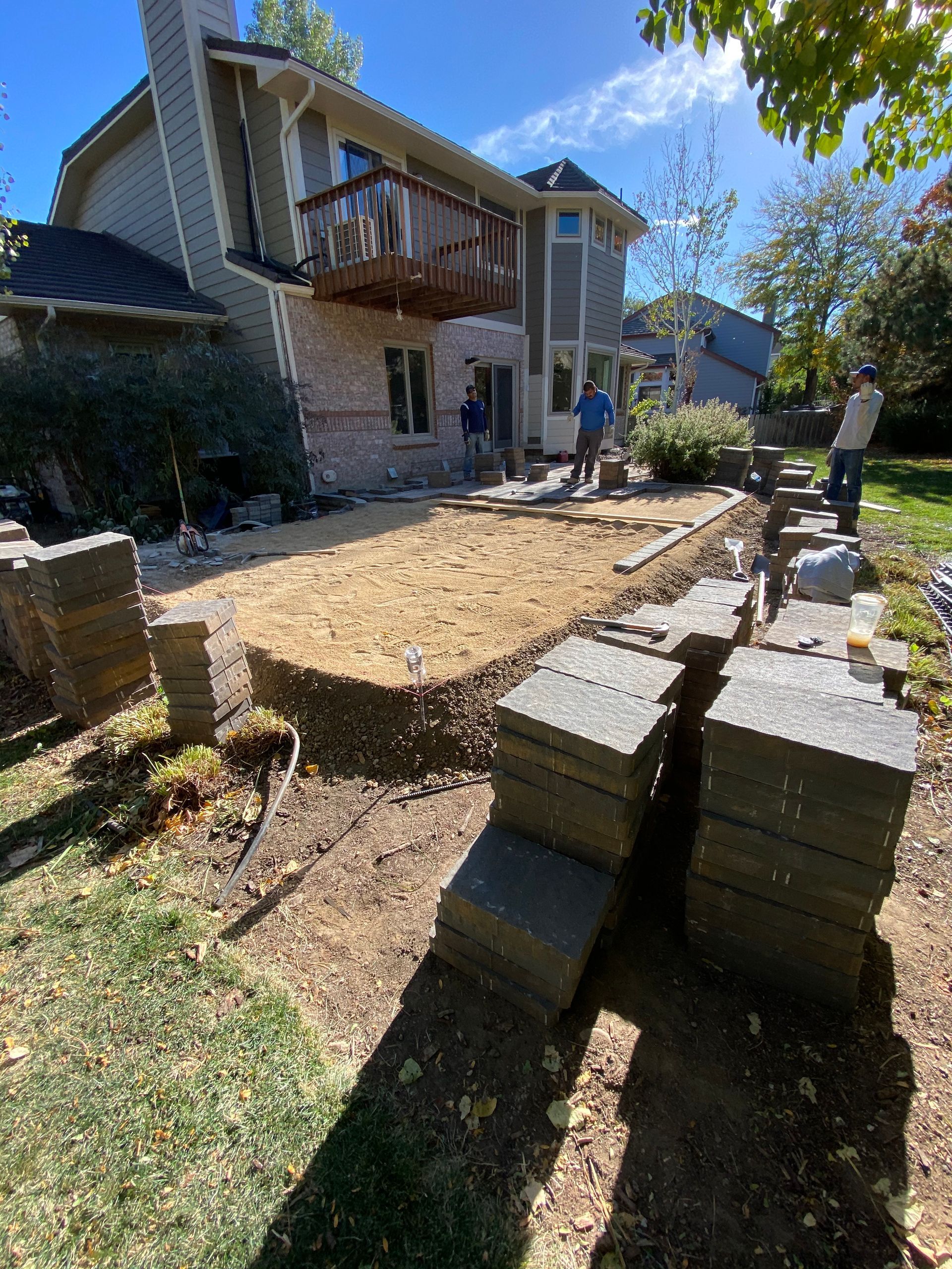 Backyard patio construction: Workers install paving stones near a house with a deck.