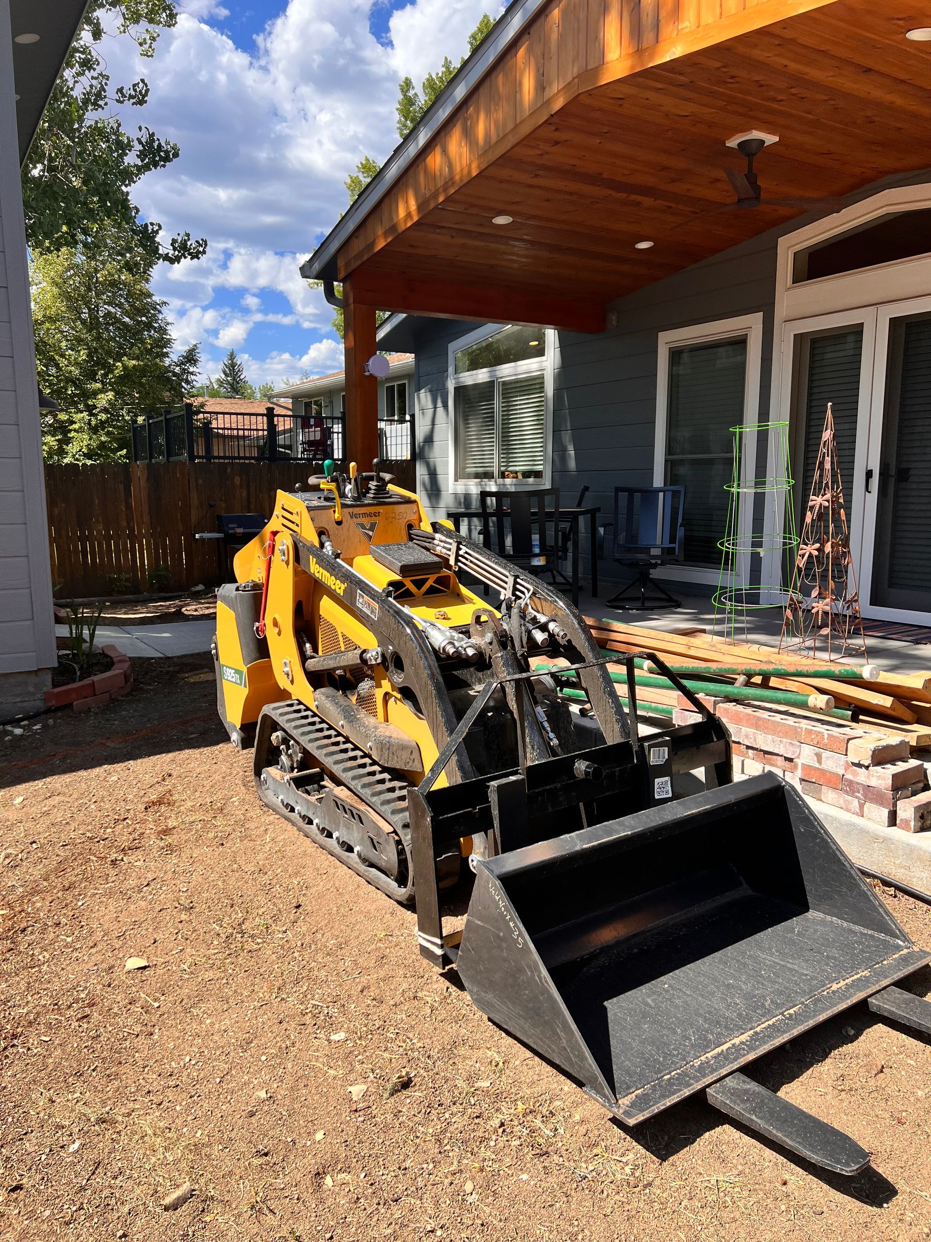 Yellow skid steer with bucket and forks on a gravel surface near a house with a porch.