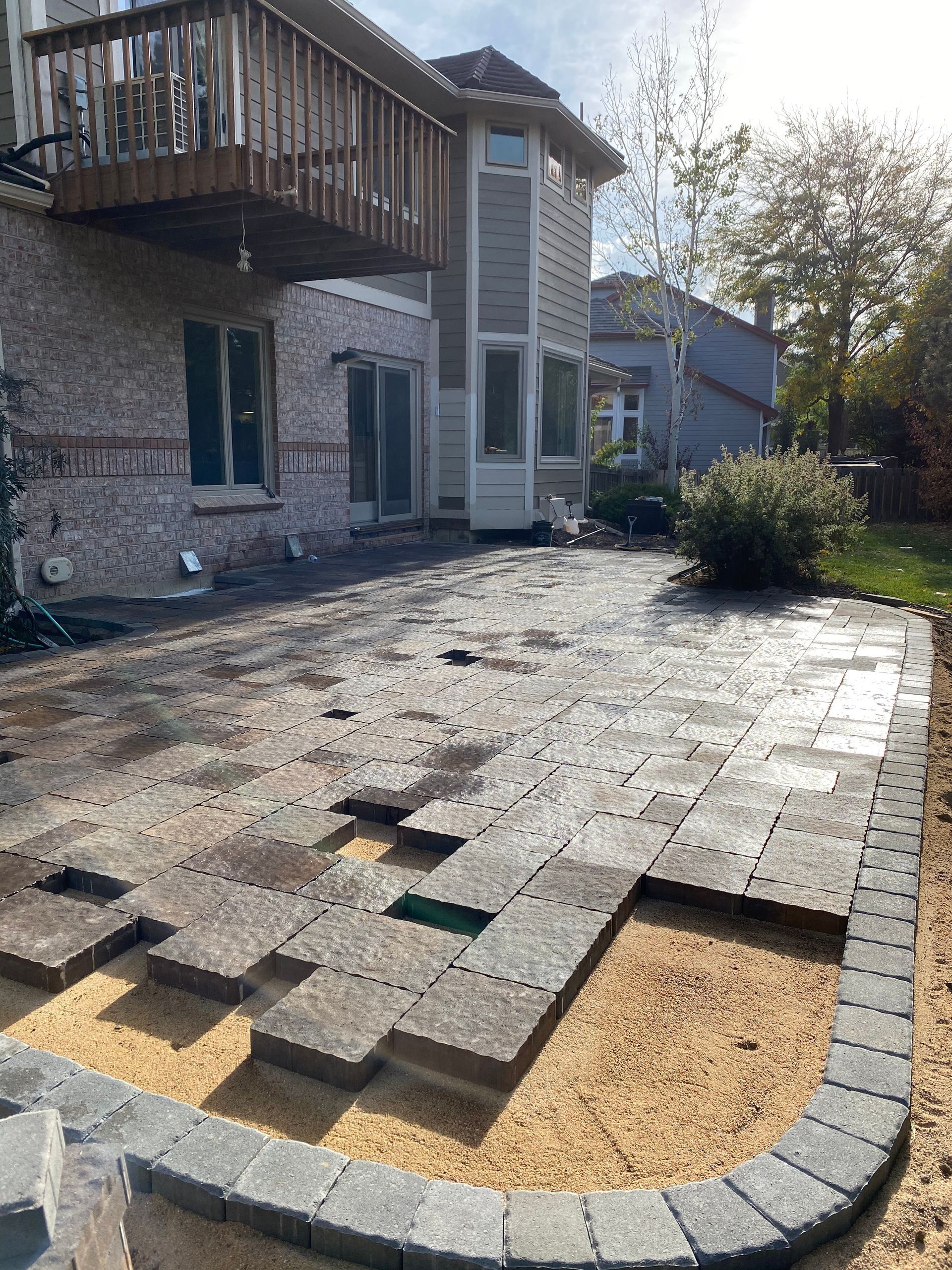 Patio construction: grey pavers laid on sand, bordered by dark stone, next to a multi-story house.