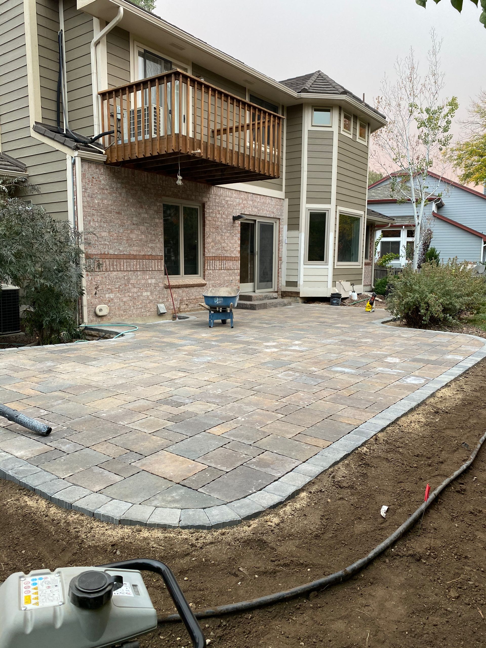 Backyard patio construction: pavers laid, edging installed, next to a two-story house with a wooden deck.