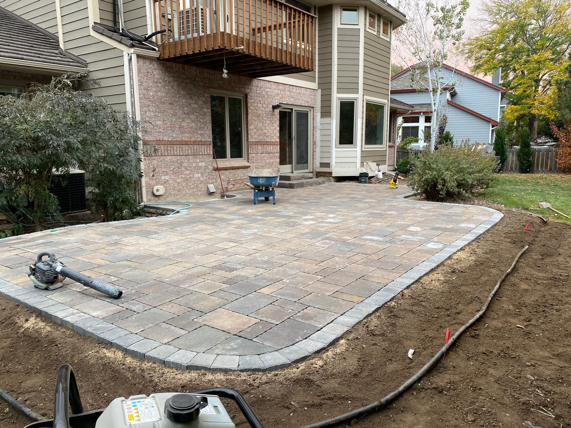 Backyard patio construction with brick pavers and edging, near a house with a balcony.