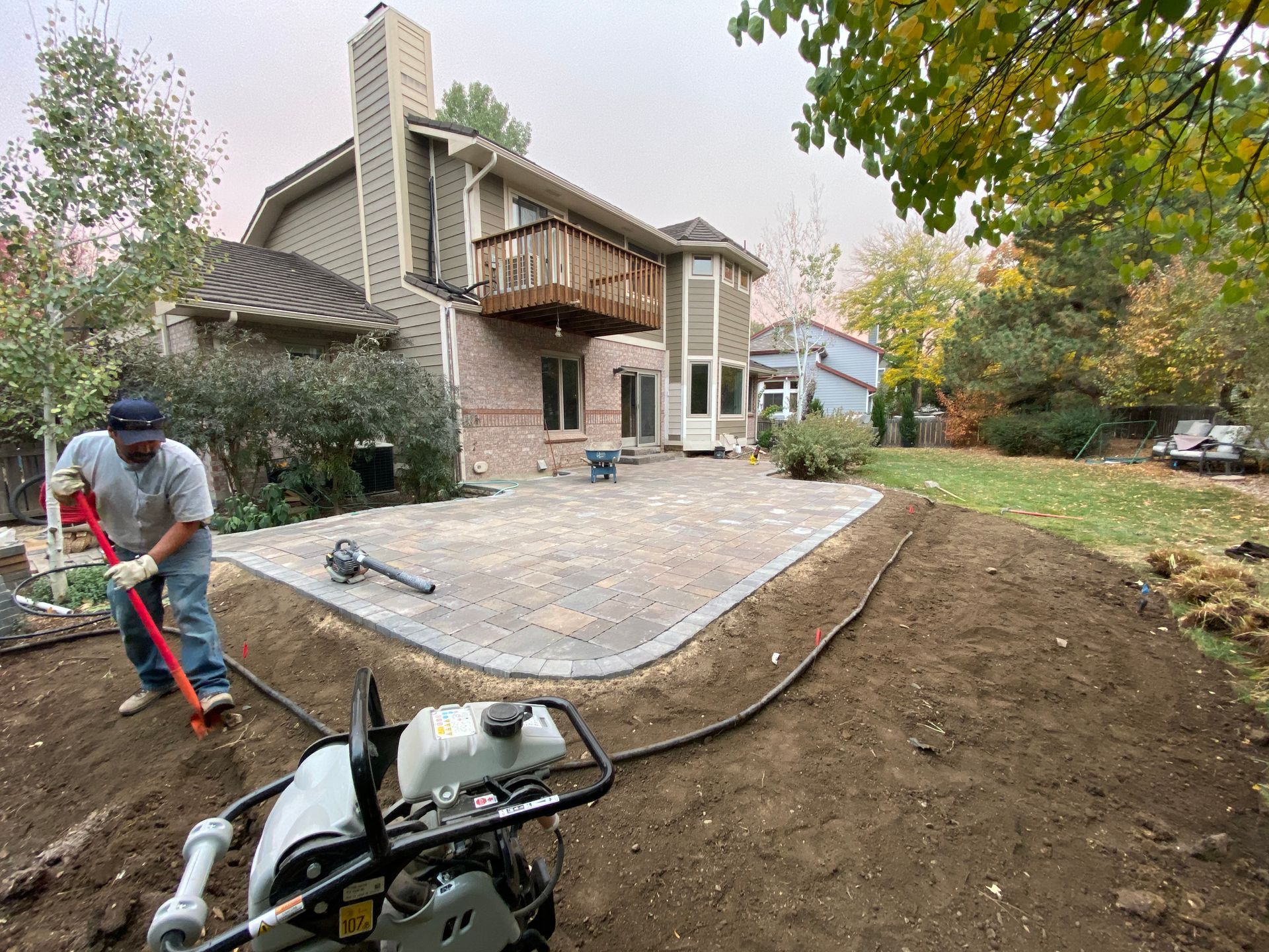 Man shovels dirt beside a new brick patio under construction near a two-story house with a wooden deck.