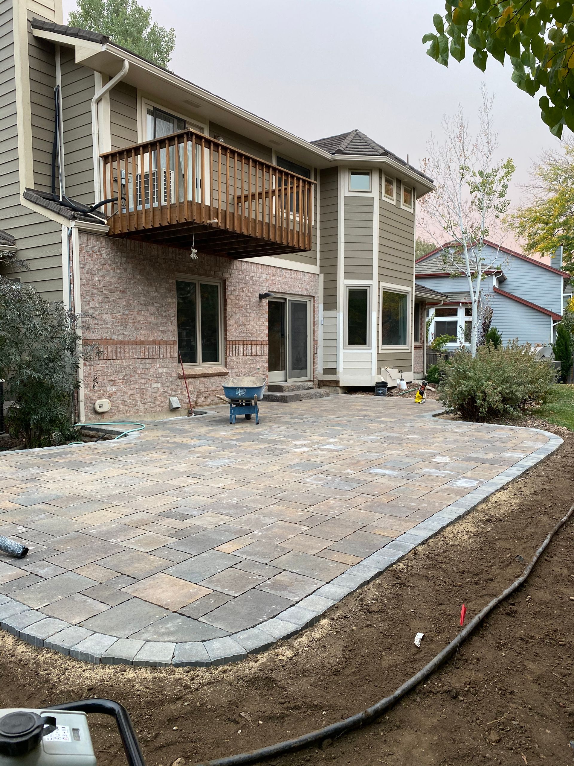 Backyard patio construction with brick house. Gray pavers being laid, surrounded by dirt.