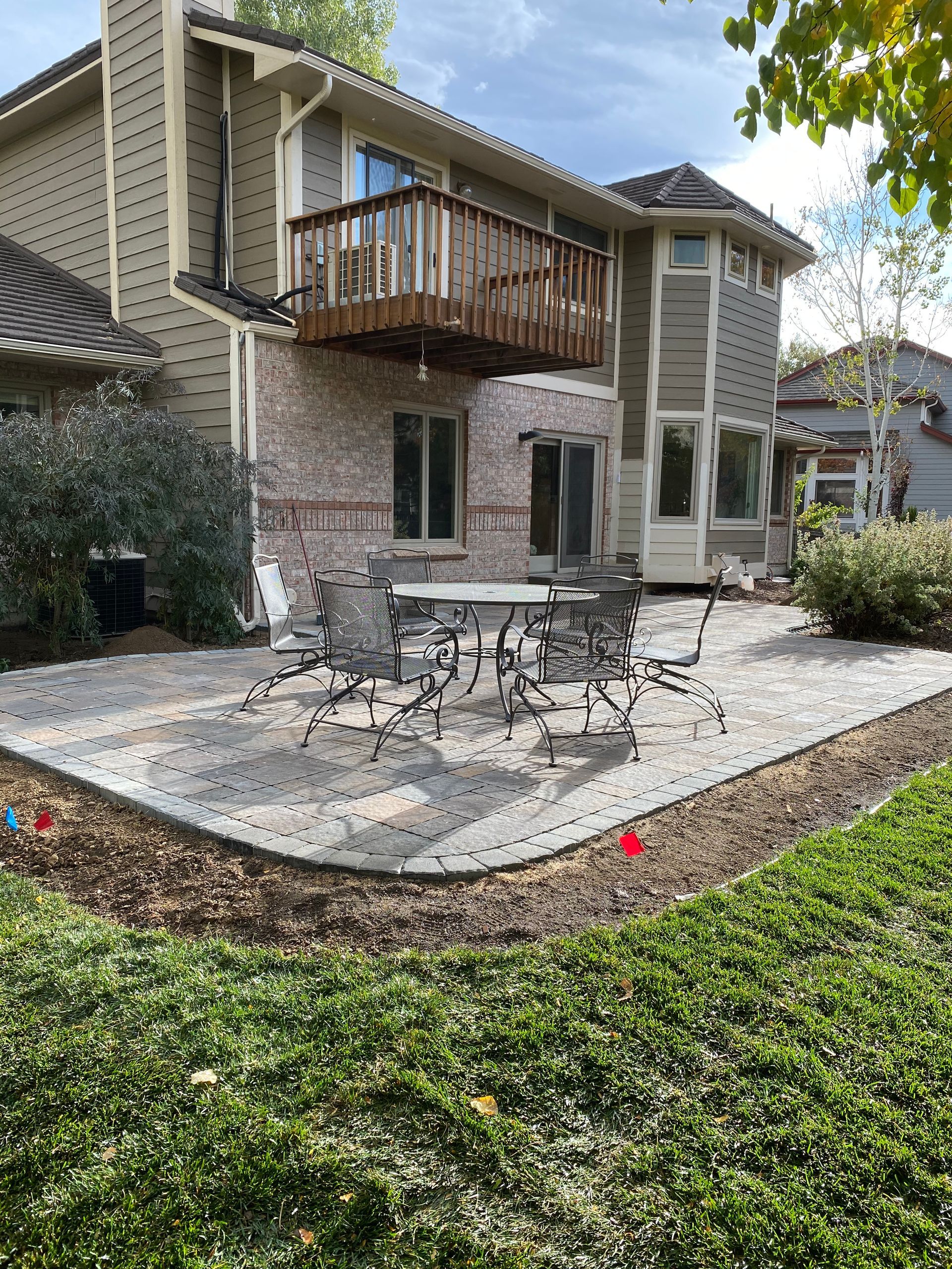 Back patio with outdoor furniture, edged with stone and bordered by greenery, adjacent to a multi-story home.