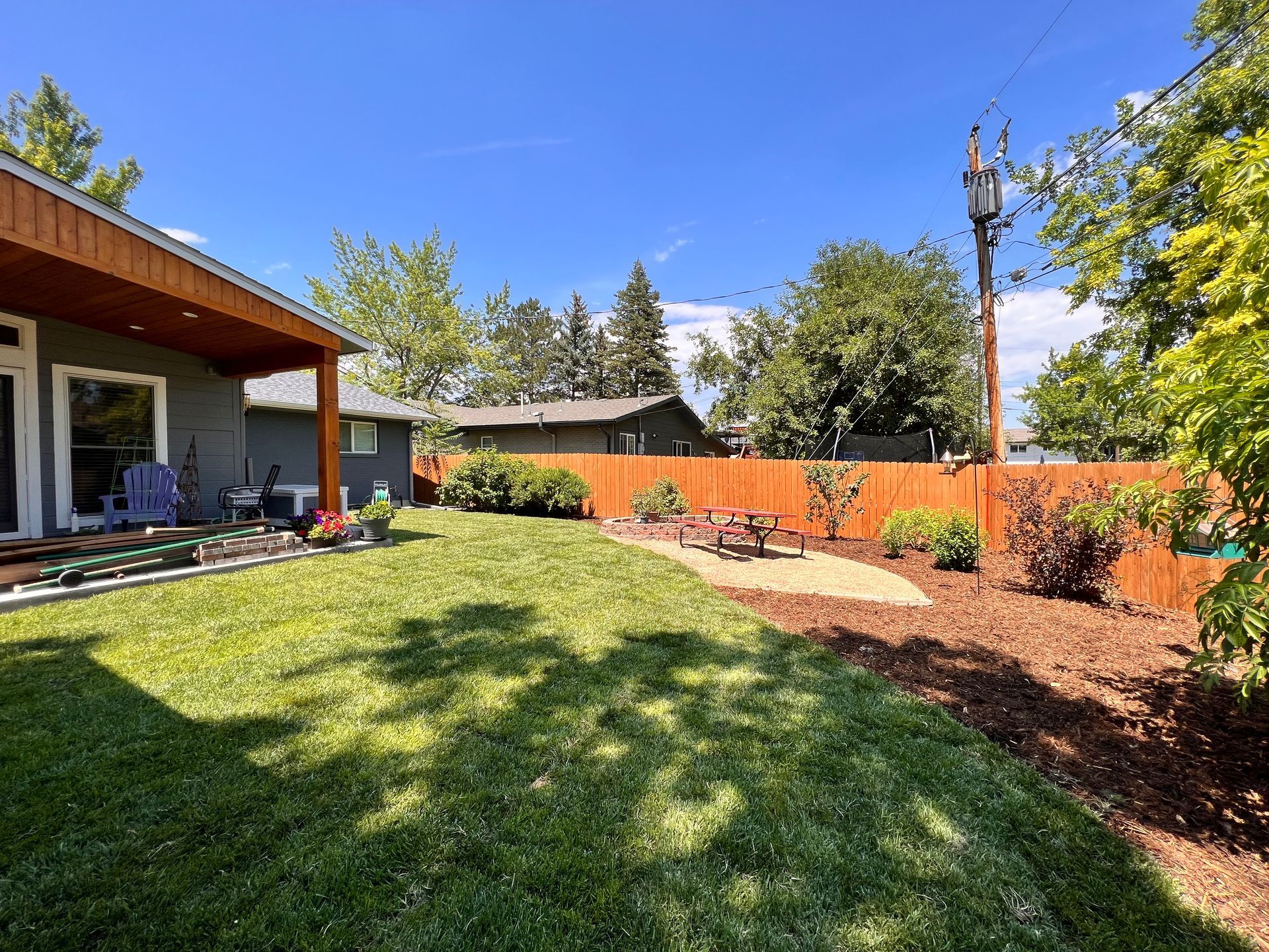 A sunny backyard with green grass, a red fence, and a patio with a picnic table.