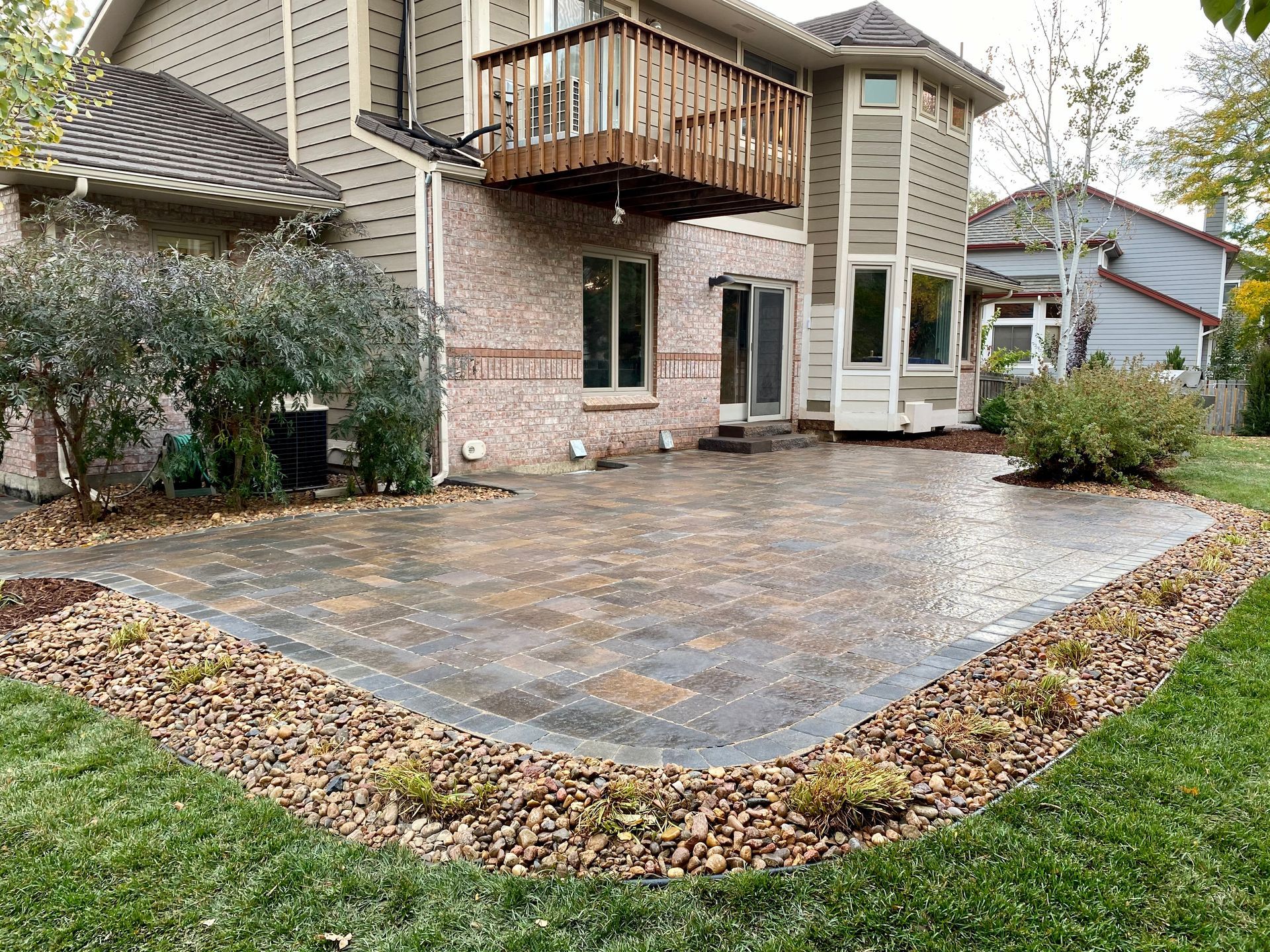 Backyard patio with stone pavers, bordered by rocks and greenery, adjacent to a two-story house with a wooden deck.