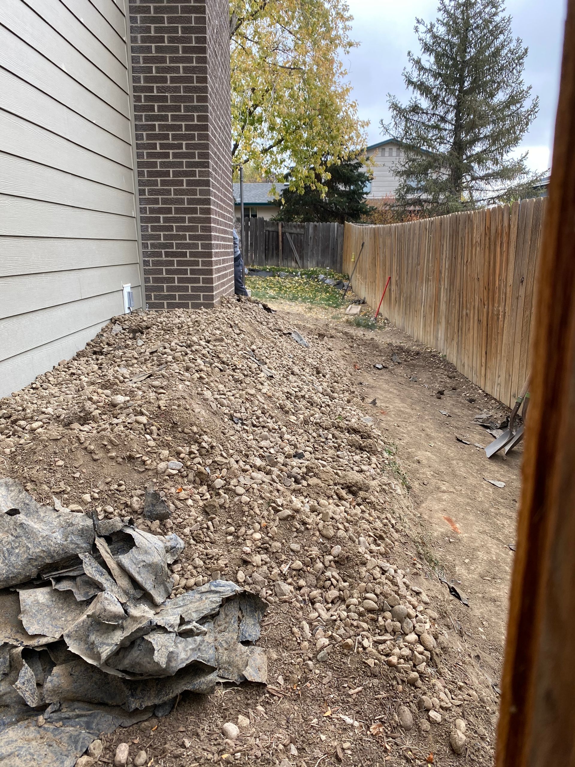 A narrow backyard path next to a fence and house, covered in gravel and dirt.