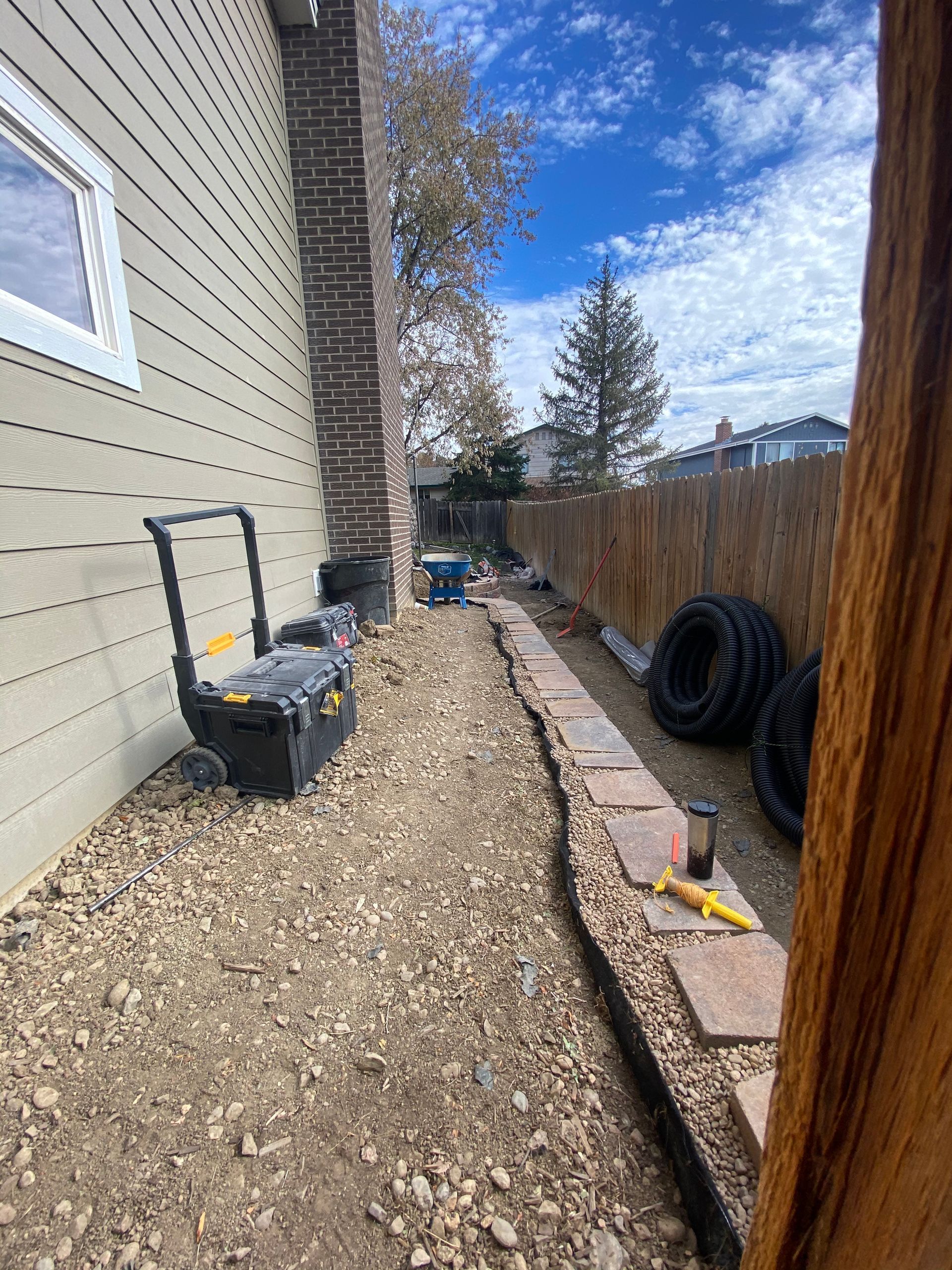 Narrow gravel path next to a house with a stone border, tools, and a wooden fence.
