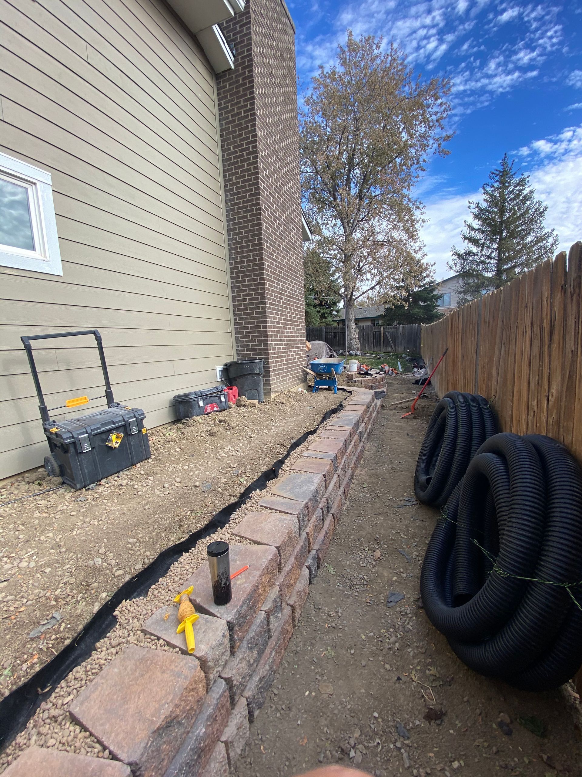 Backyard construction scene: brick retaining wall, black drainage pipes, gravel, tools, brown fence, and a chimney.