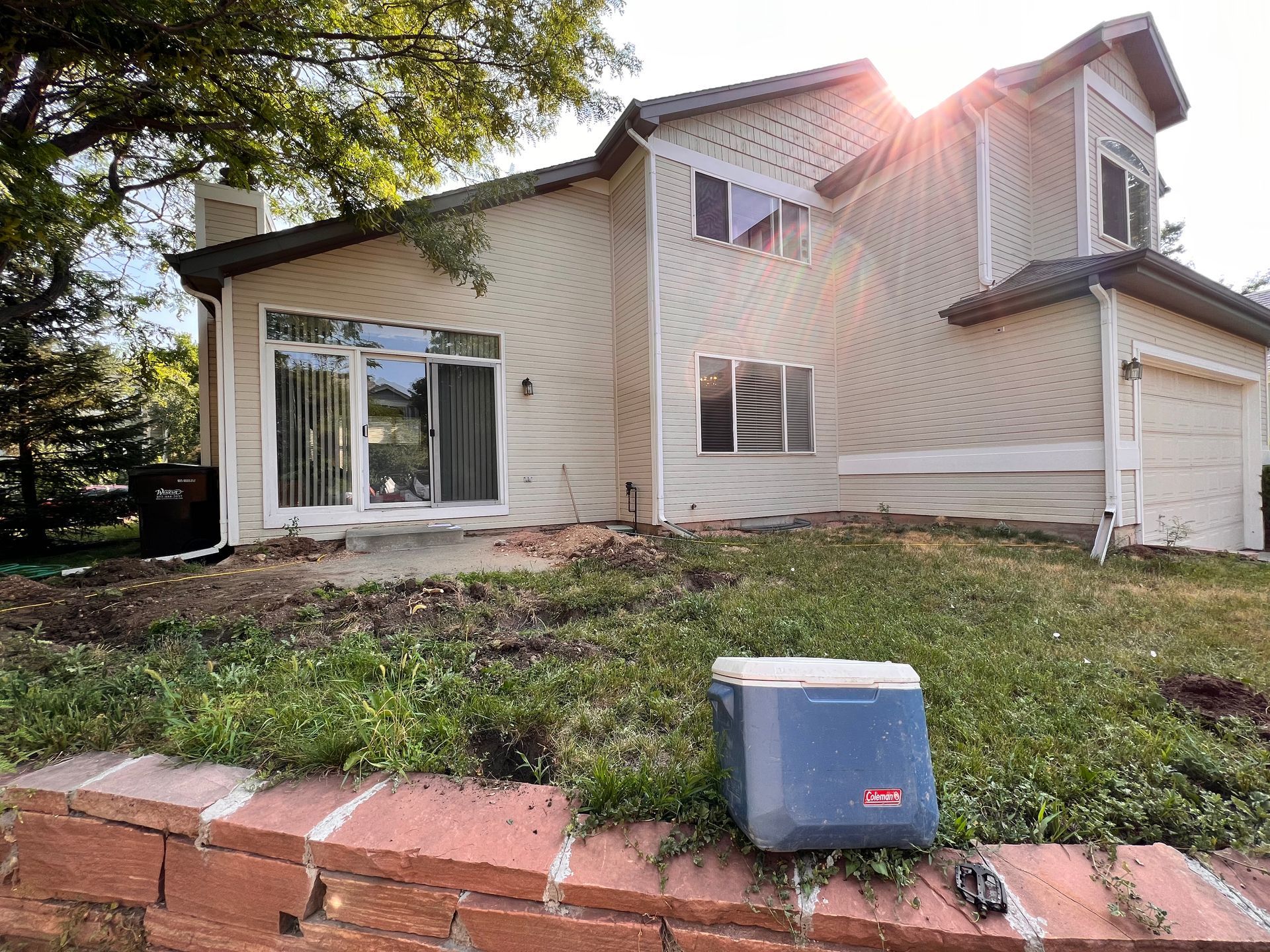 Two-story beige house with sliding glass door and garage, blue cooler on a brick retaining wall.