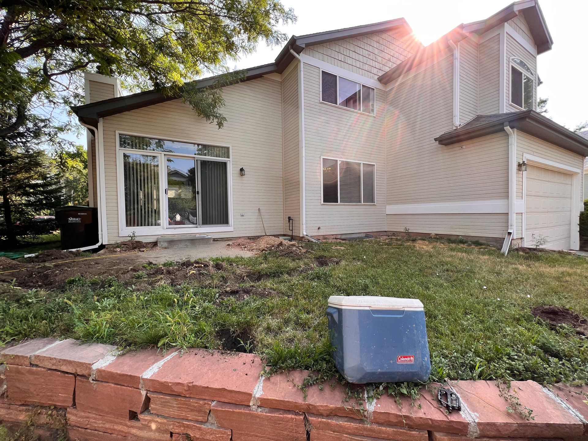 Beige two-story house with a brick wall and a blue cooler on a grassy hill. Sun shining.