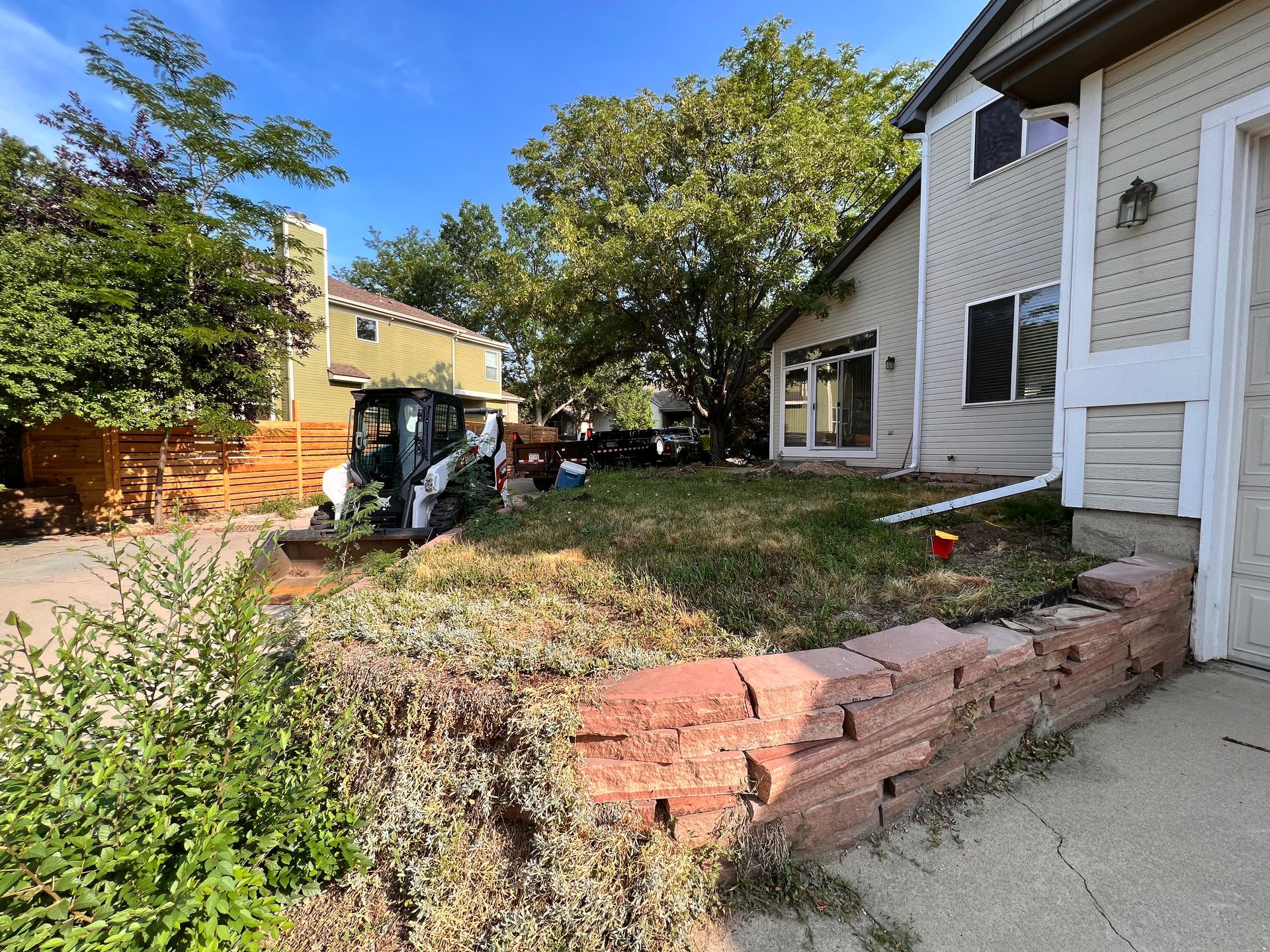 A small Bobcat machine in a yard, a brick retaining wall and two-story house.