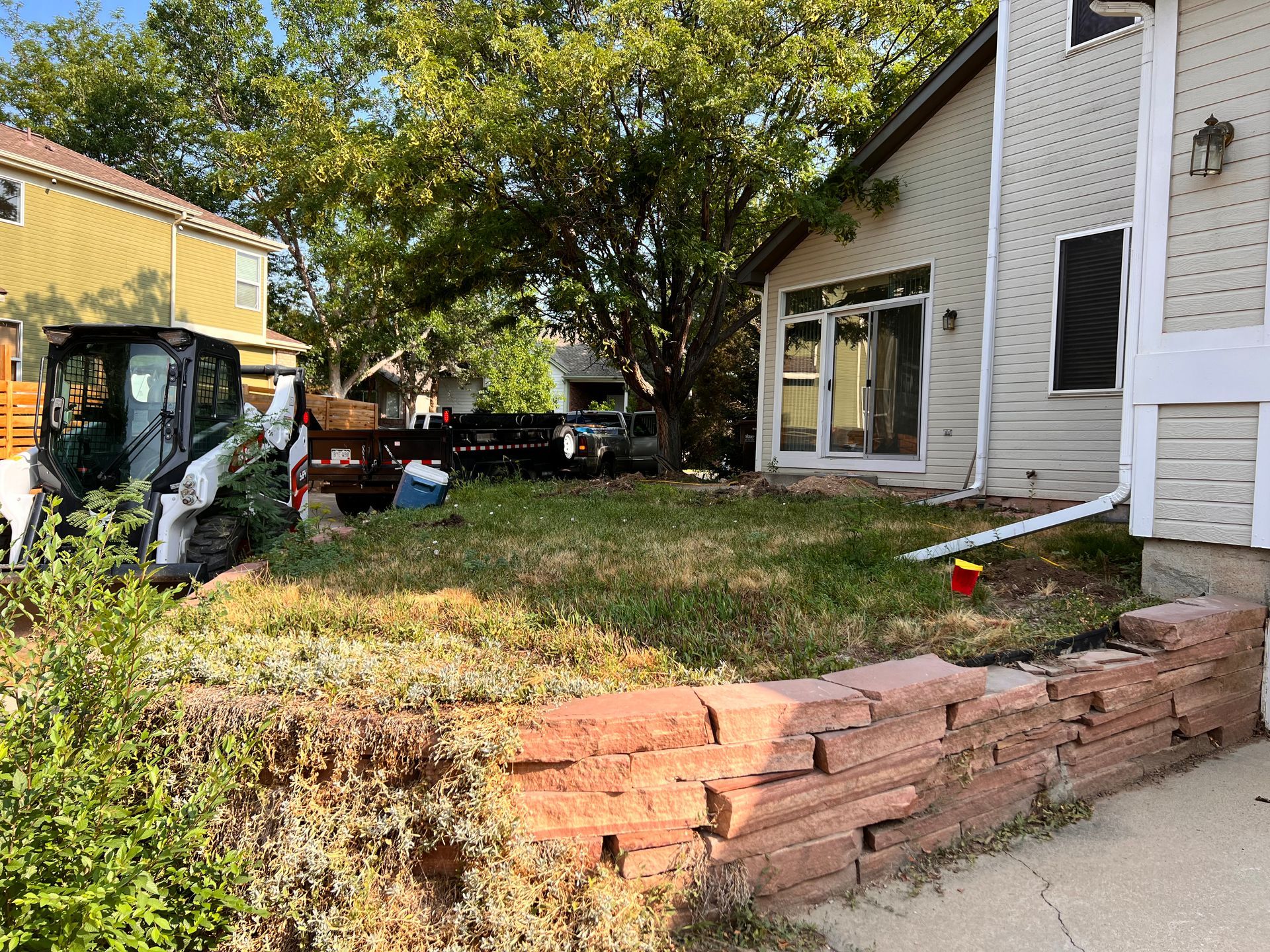 Bobcat near a house with red brick retaining wall and overgrown grass.