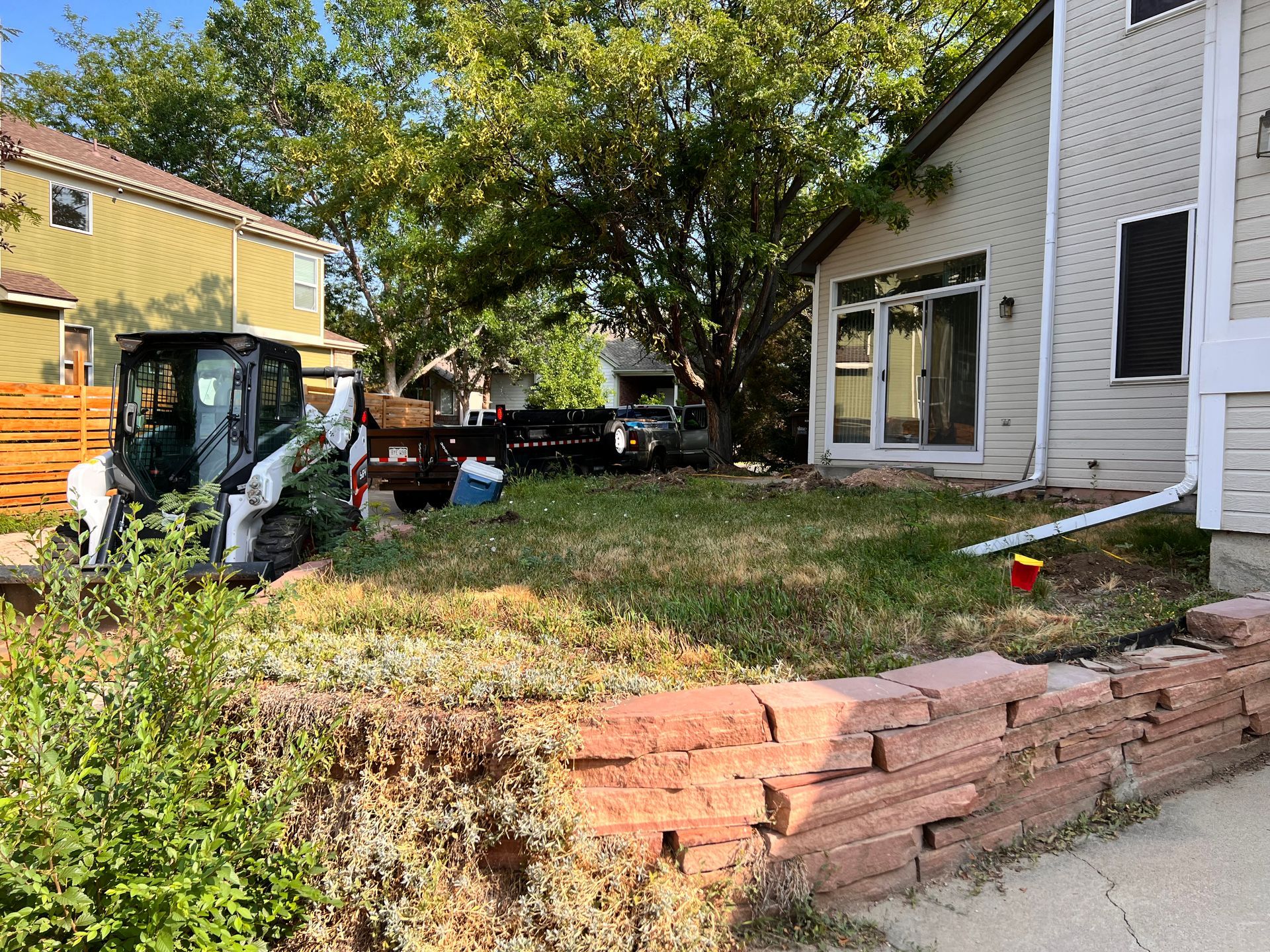 Bobcat, trailer, and house in a grassy yard, with a brick retaining wall in the foreground.