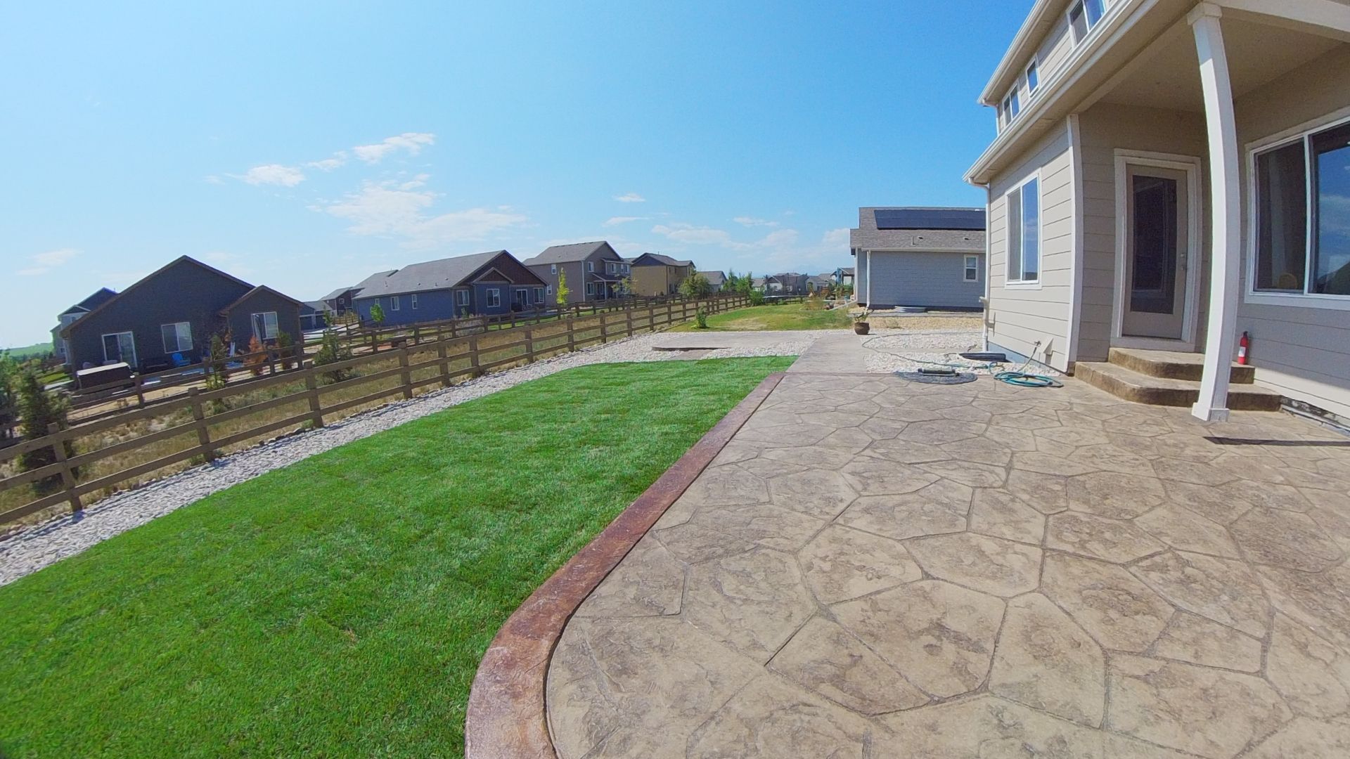 Backyard with a stamped concrete patio, green lawn, wooden fence, and beige house on a sunny day.