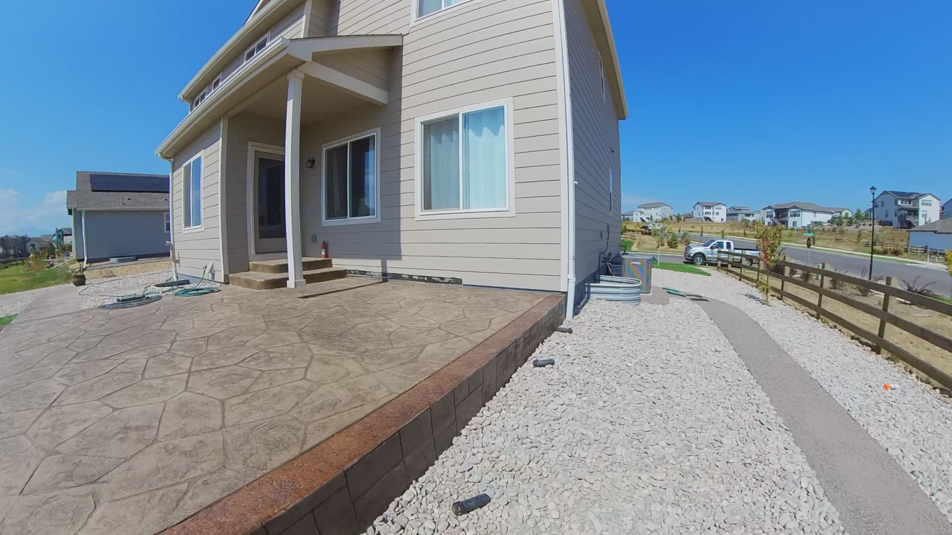 Two-story beige house with a concrete patio, gravel pathway, and a fence under a bright blue sky.