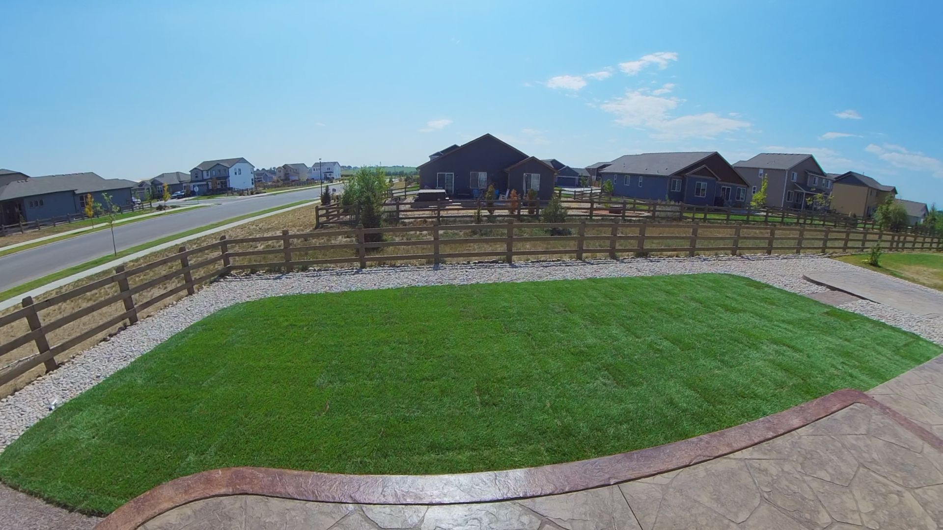 Green lawn in a backyard, bordered by a fence and a decorative stone path, houses in the background under a blue sky.