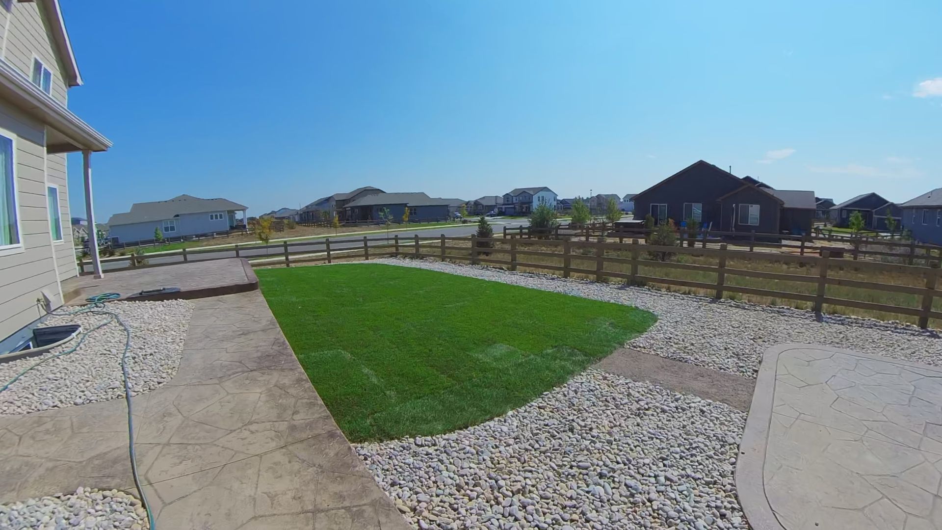 Lawn in a residential backyard, fenced, with gravel path and surrounding houses under a blue sky.