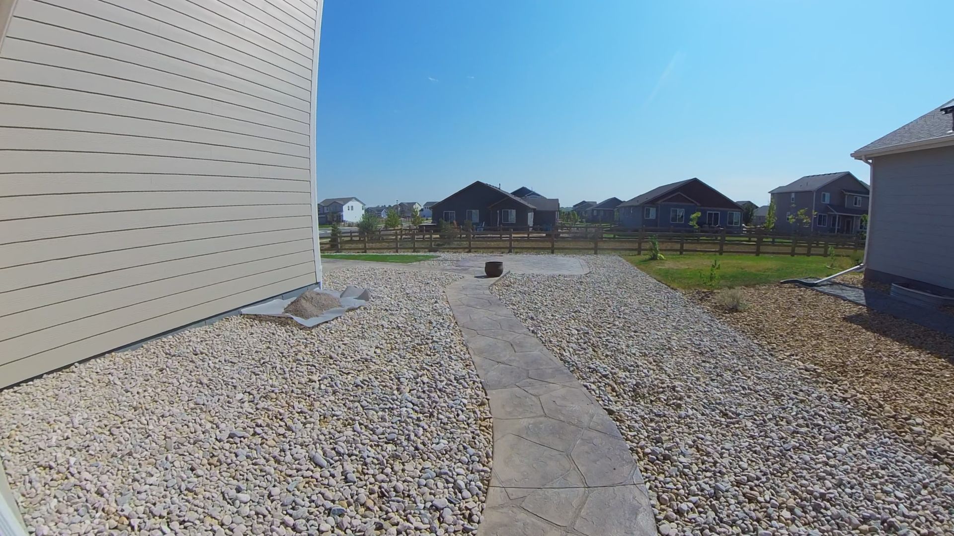 Backyard with stone path, gravel, and houses under a clear blue sky.