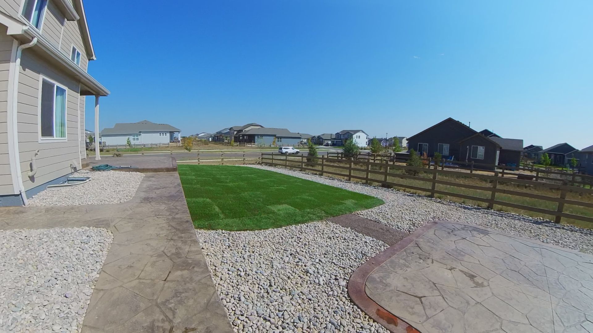 A sunny backyard with a concrete path, rocks, and a small patch of green grass; other houses in the distance.