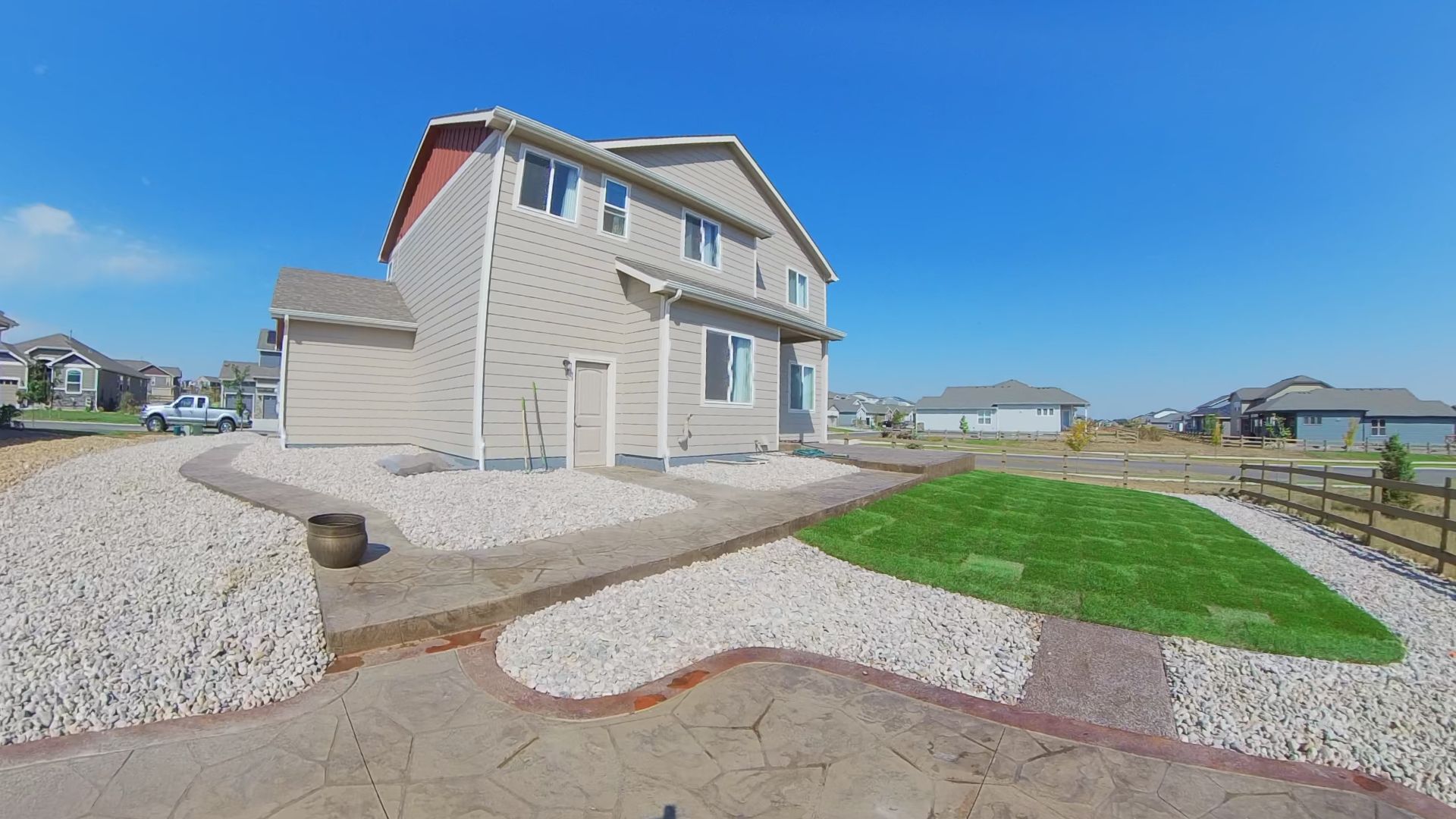 Two-story house with tan siding, red roof, gravel landscaping, and green grass against a blue sky.