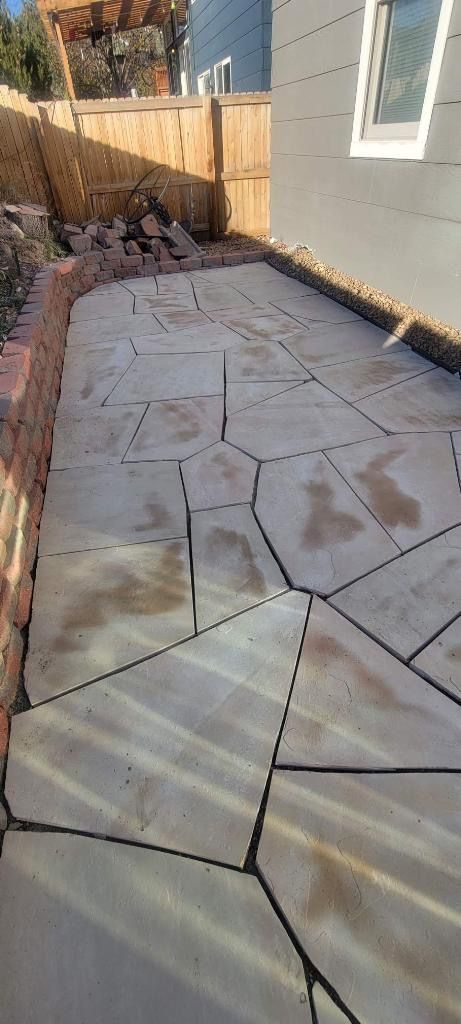 Stone patio next to a house with a wooden fence and retaining wall in the background.