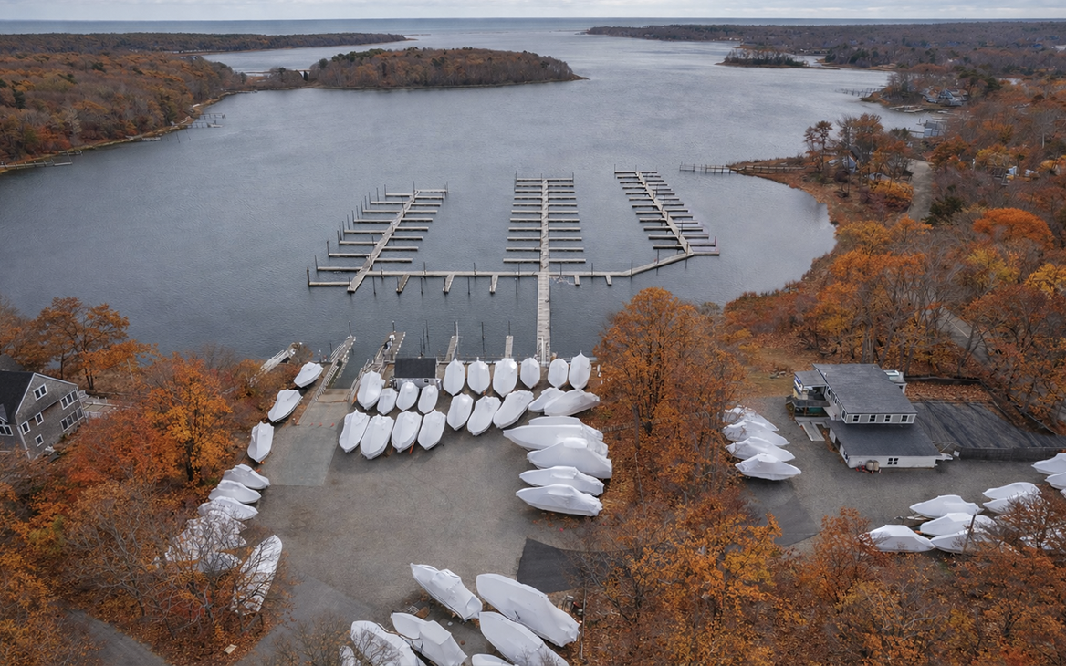 Aerial view of a marina with boat slips and covered sailboats, surrounded by autumn trees and a bay.