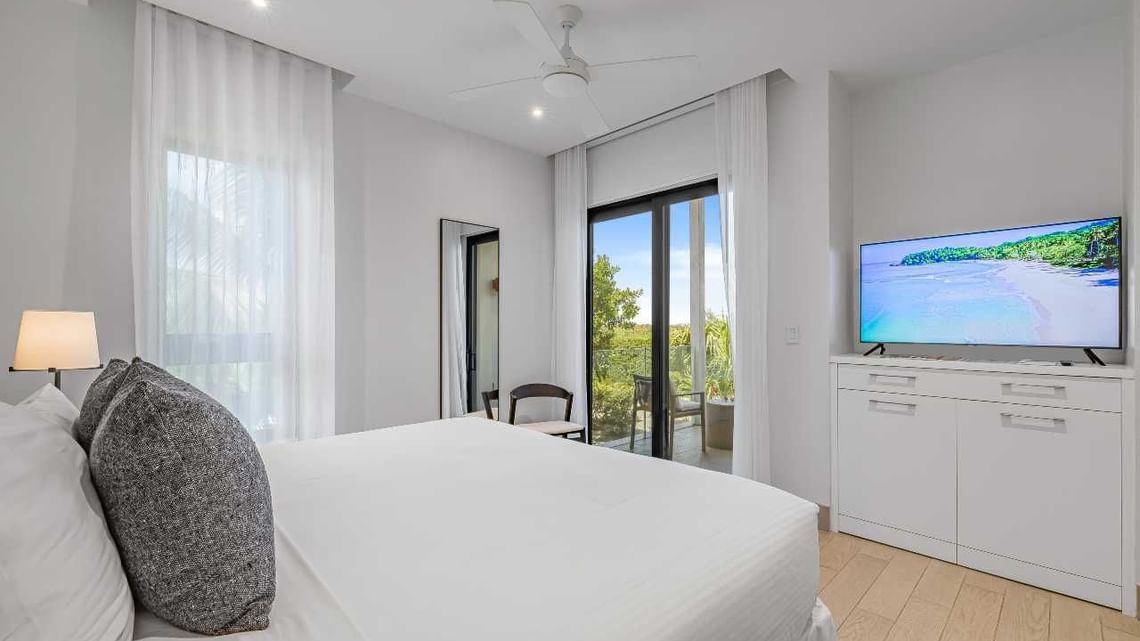 Bright modern bedroom with white bed, floor-to-ceiling curtains, and TV on a cabinet near a sliding door.