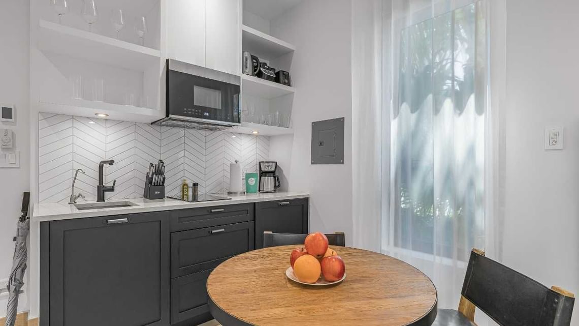 Bright kitchen with gray cabinets, white countertops, and a round table with fruit near a window.