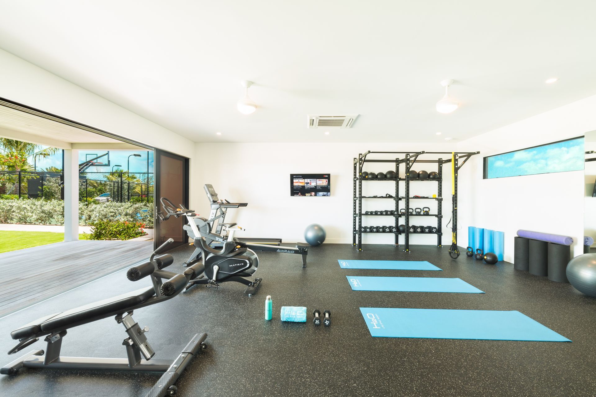 Bright fitness studio with mats, exercise bikes, weights, and shelves, lit by large windows