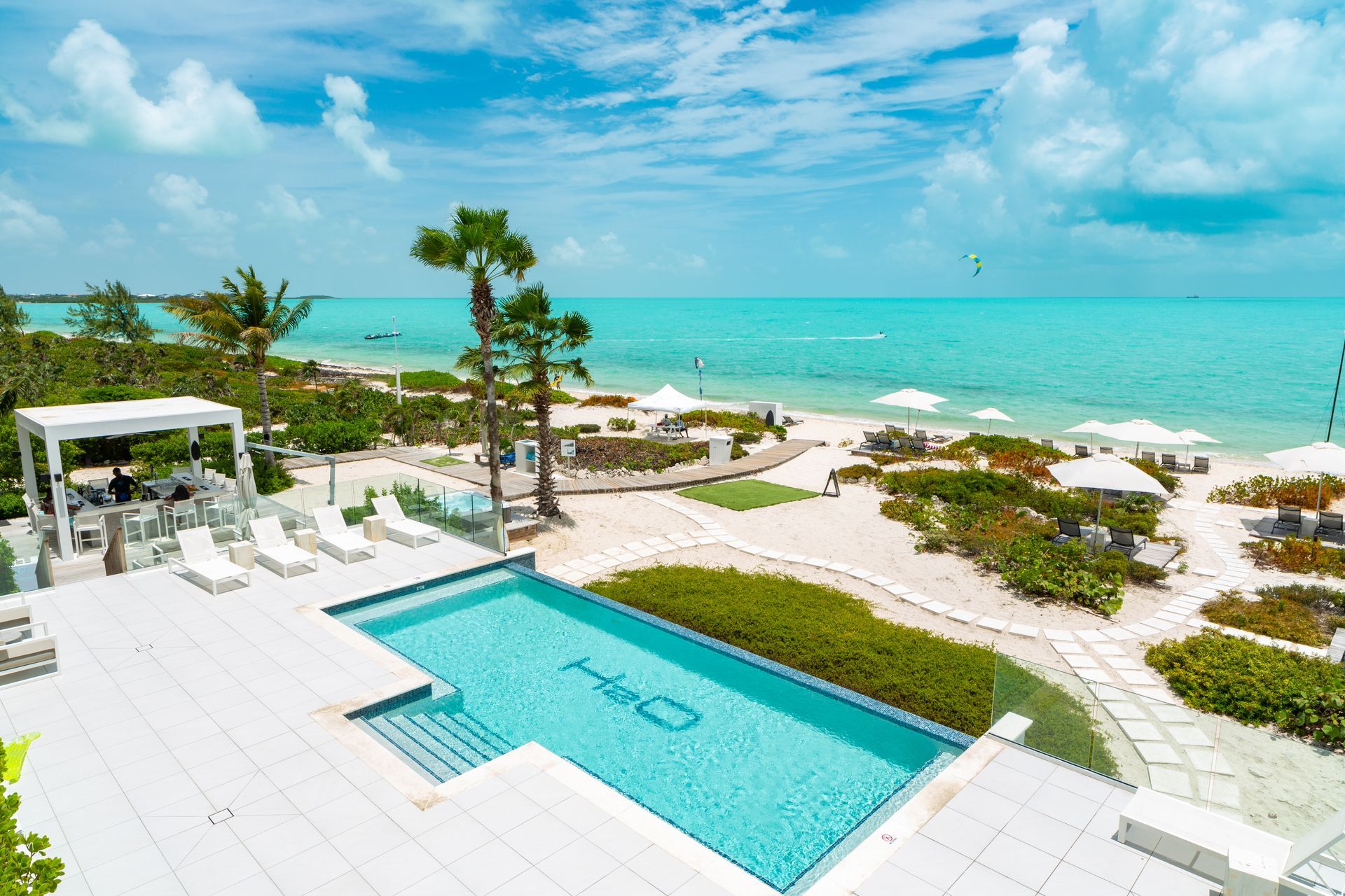 Beachfront resort with turquoise pool, white lounge chairs, and ocean view under a bright blue sky
