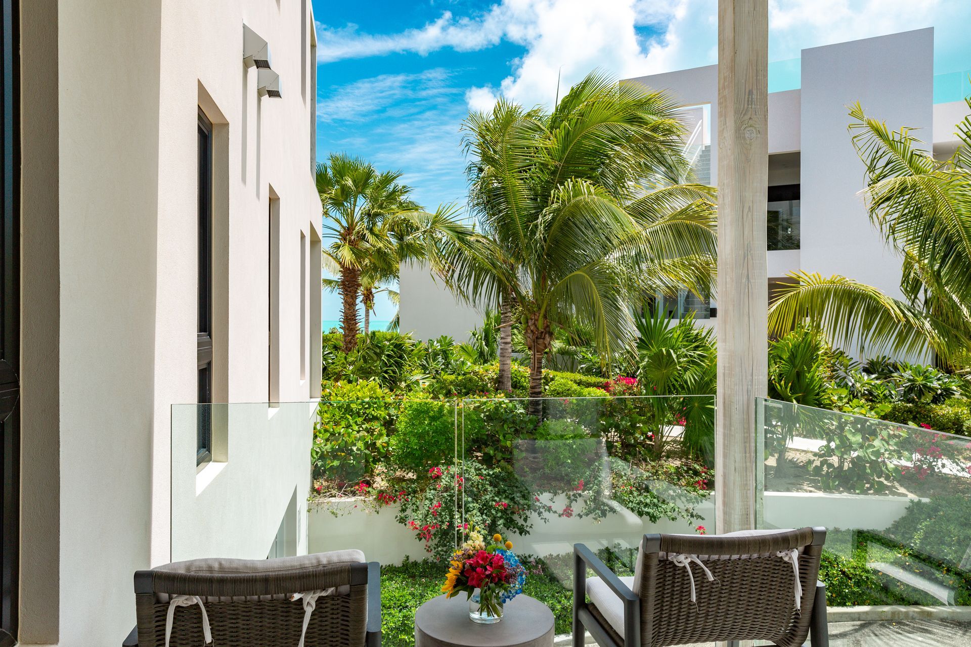 Balcony with palm trees, glass railing, and chairs overlooking a bright tropical garden
