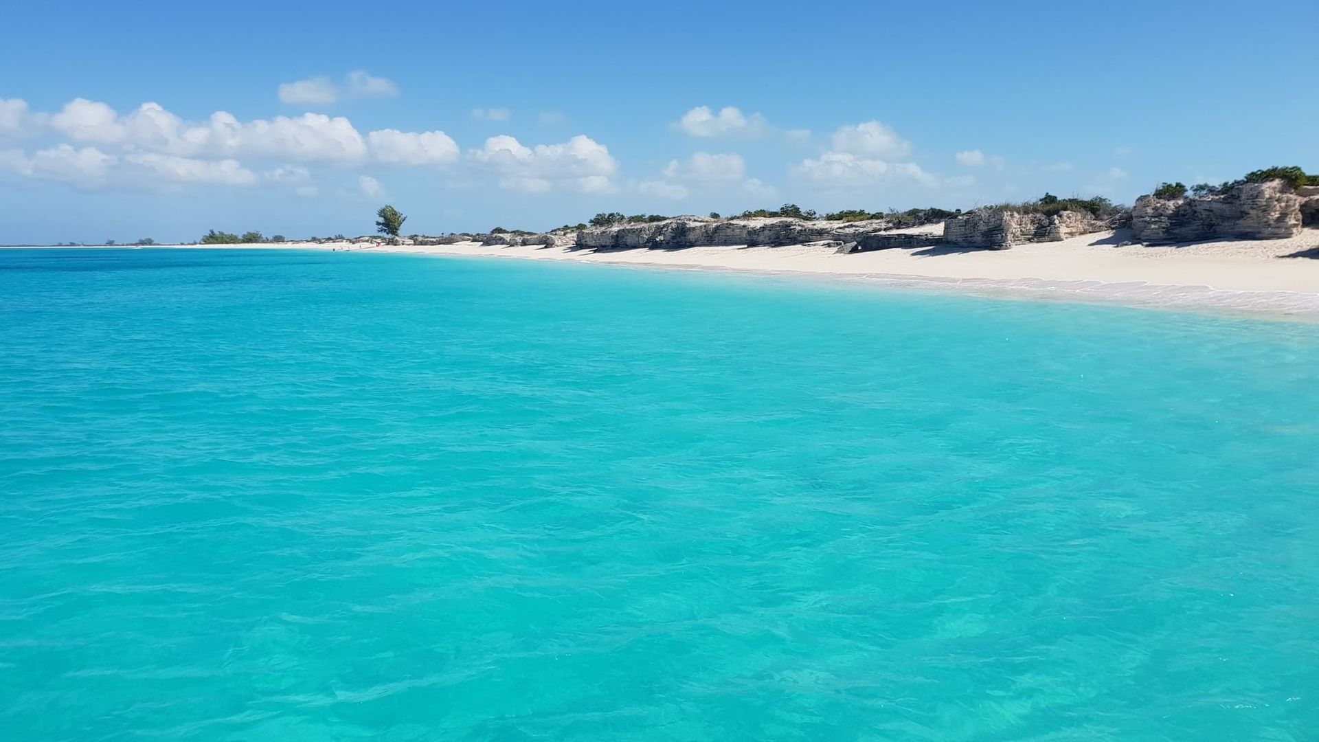Turquoise sea beside a sandy beach under a blue sky, with distant rocks and houses.