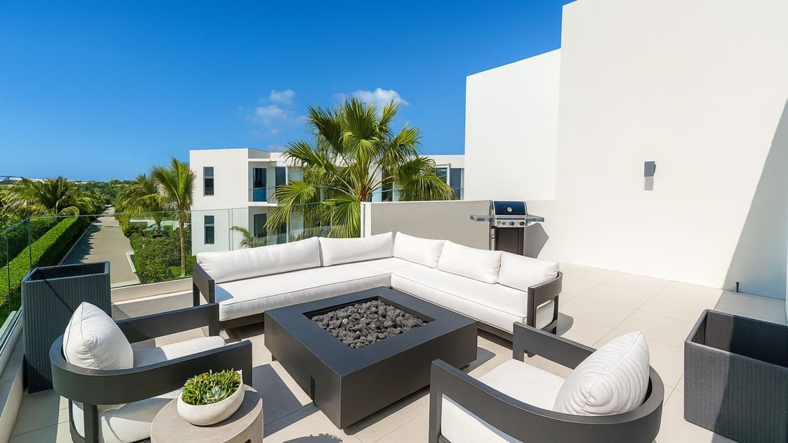 Modern rooftop patio with gray sofas, fire pit, and palm trees by white buildings under a blue sky