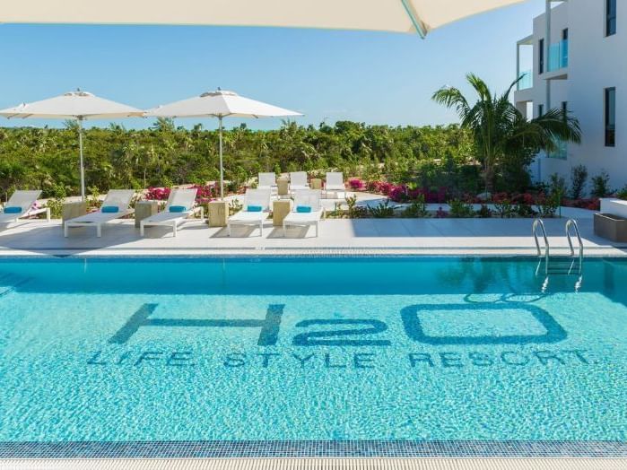 Resort pool with lounge chairs, umbrellas, and turquoise water beside a modern hotel.