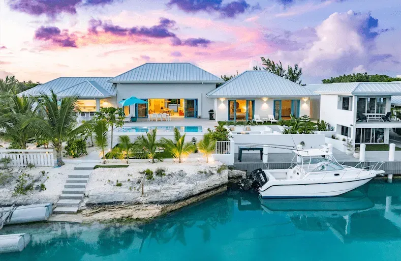 Luxury waterfront villa with pool, palm trees, and a white yacht at sunset