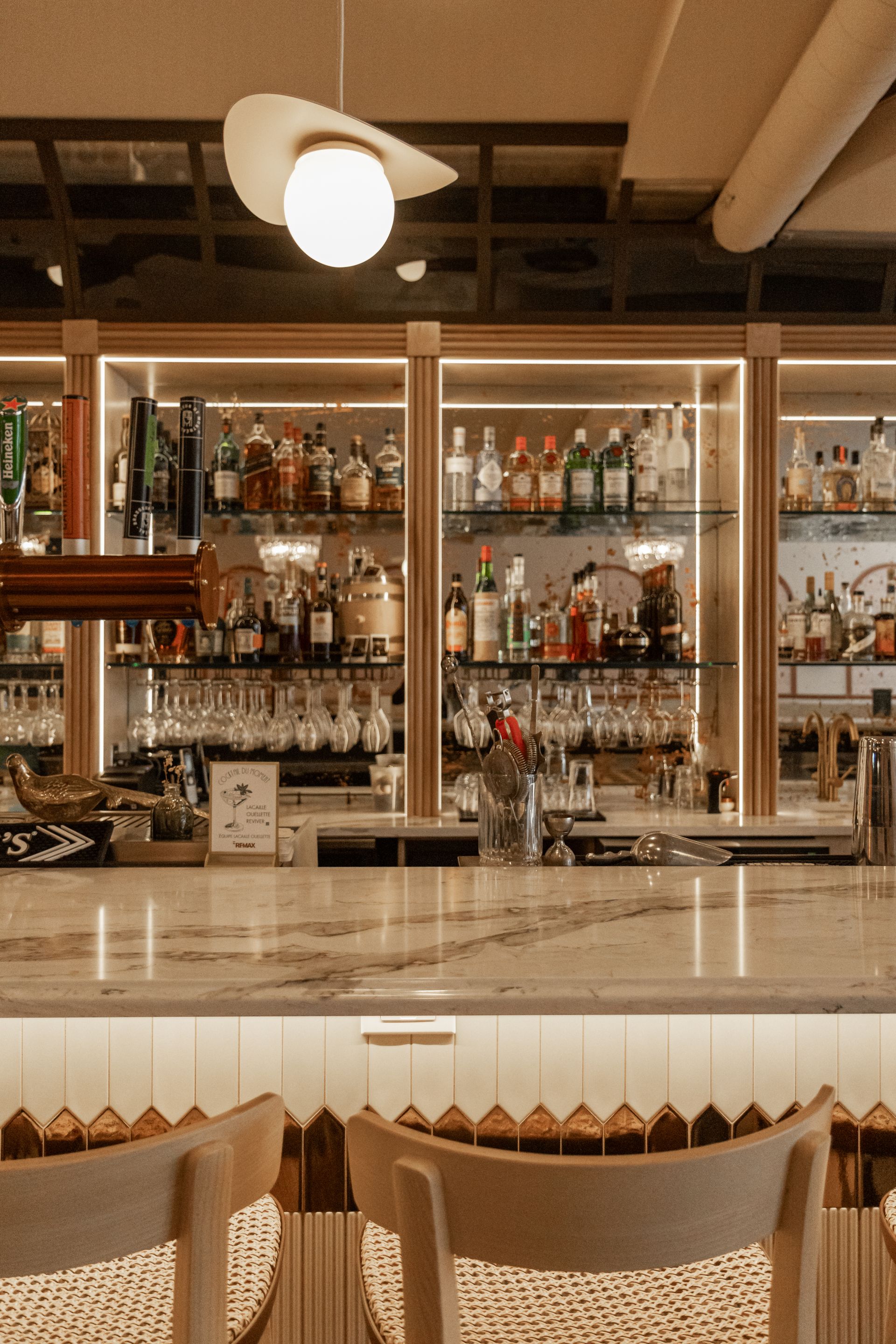 Bar chic vide avec étagères à spiritueux rétroéclairées, comptoir en marbre et tabourets en osier.