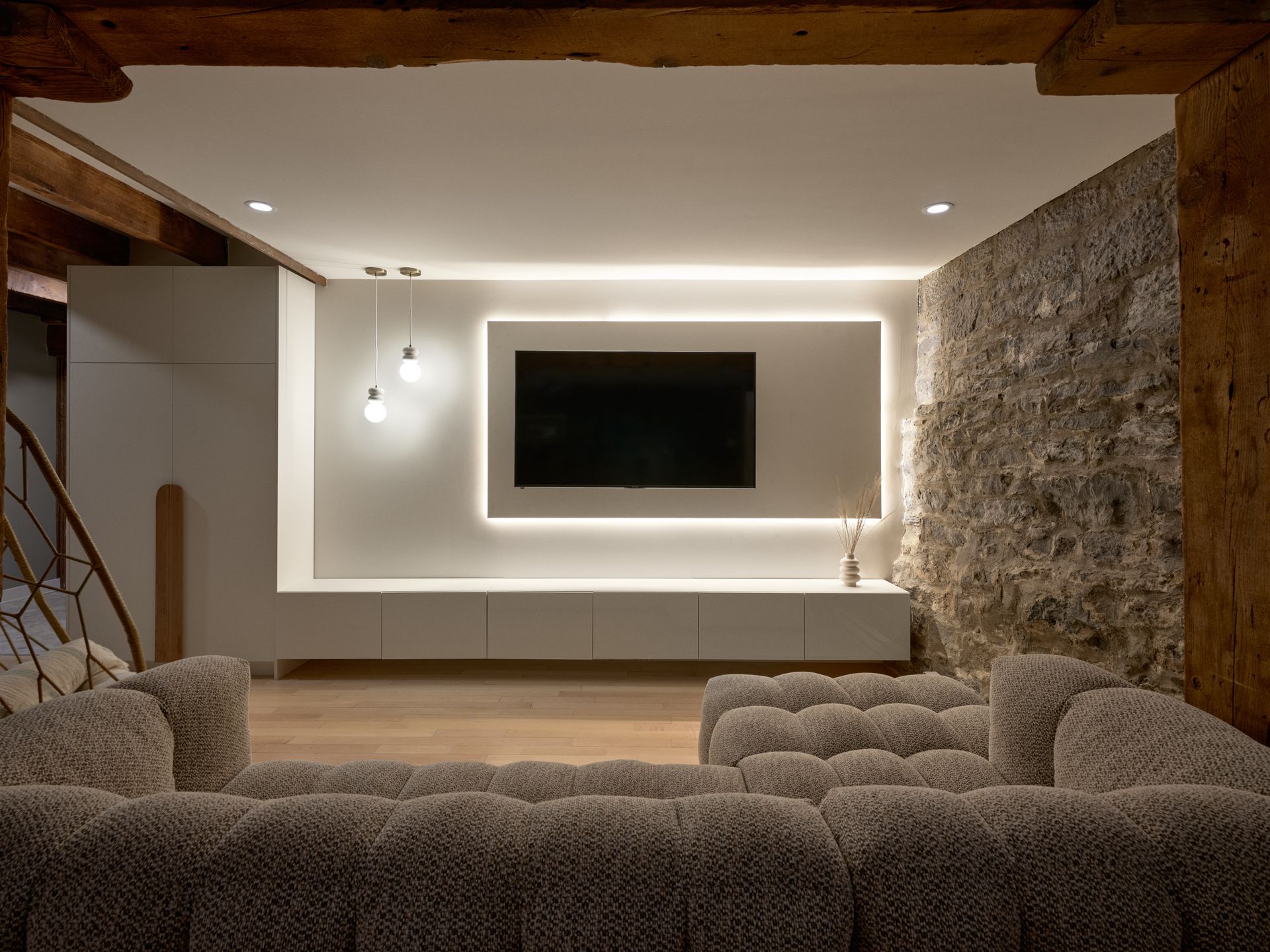 Salon avec télévision murale encadrée par un bandeau lumineux. Mur en pierre à droite, éléments encastrés blancs et canapé moelleux.