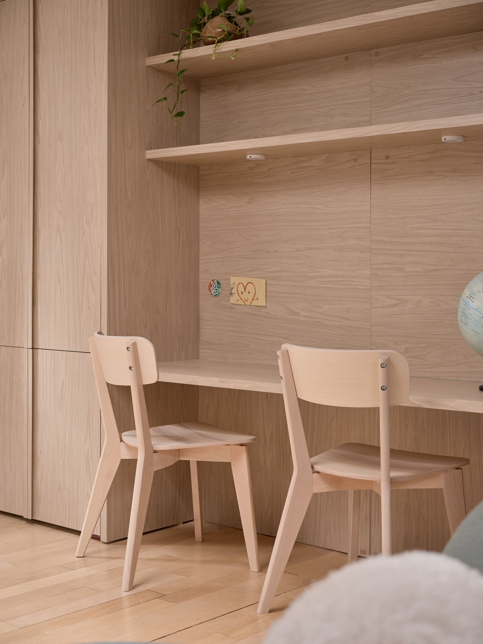 Deux chaises en bois sont disposées autour d'un bureau intégré à un meuble mural en bois muni d'étagères. Un globe terrestre et une prise électrique sont également visibles.
