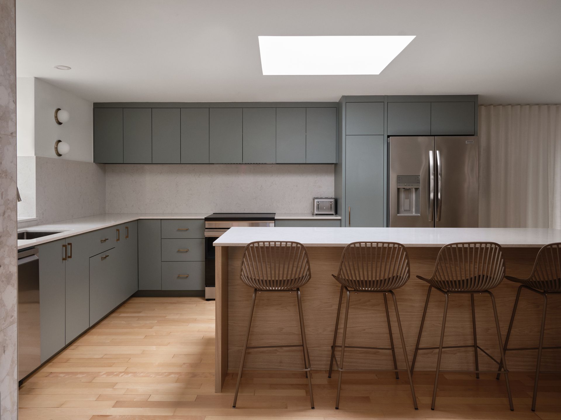 Cuisine moderne avec armoires bleu-gris, îlot en bois clair avec tabourets de bar, appareils électroménagers en acier inoxydable et puits de lumière.