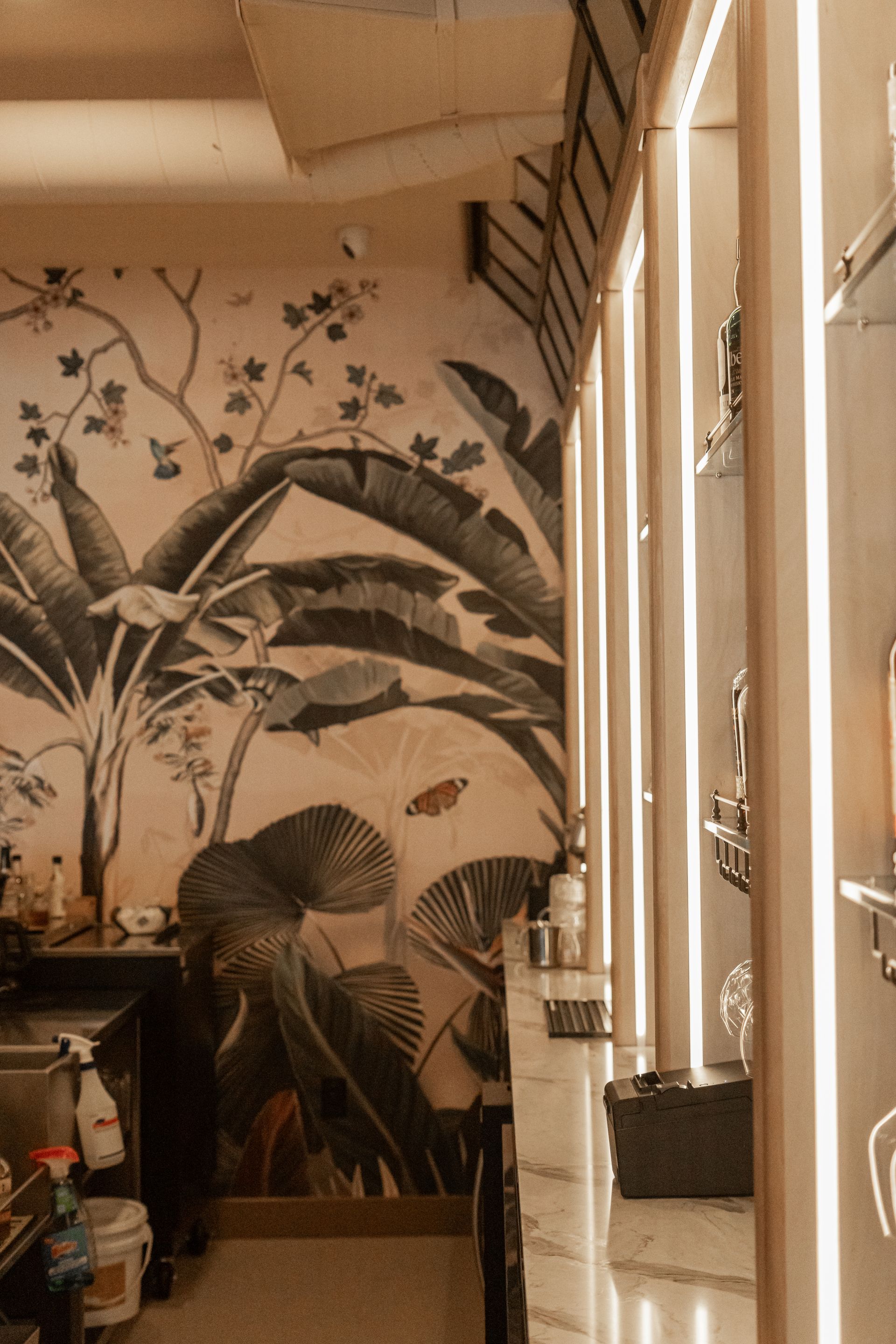 Couloir étroit orné d'une fresque tropicale, de hautes fenêtres et de plantes en pot près d'une porte lumineuse.