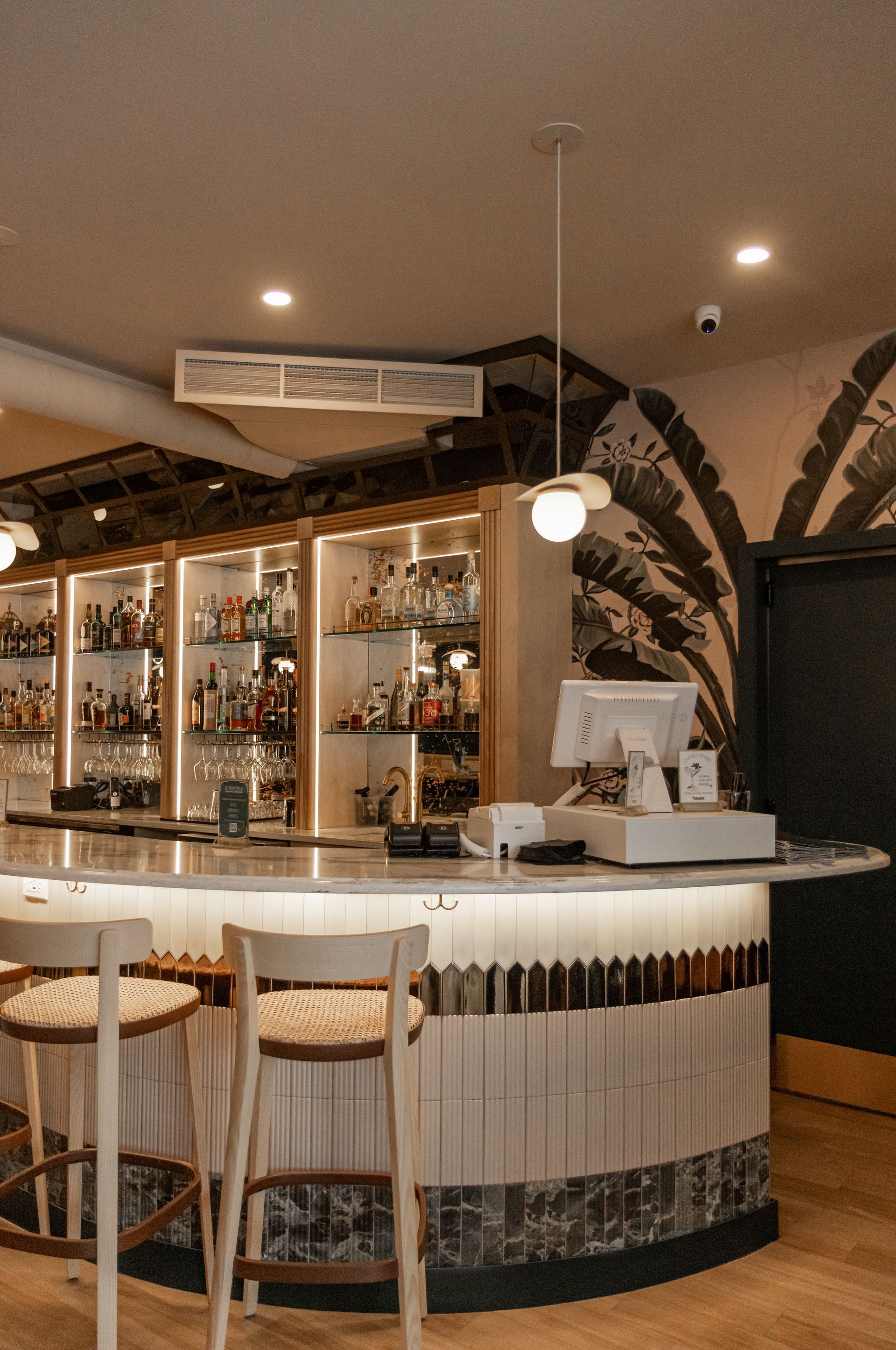 Intérieur de bar chaleureux avec comptoir blanc incurvé, étagères à bouteilles et éclairage suspendu aux tons chauds.
